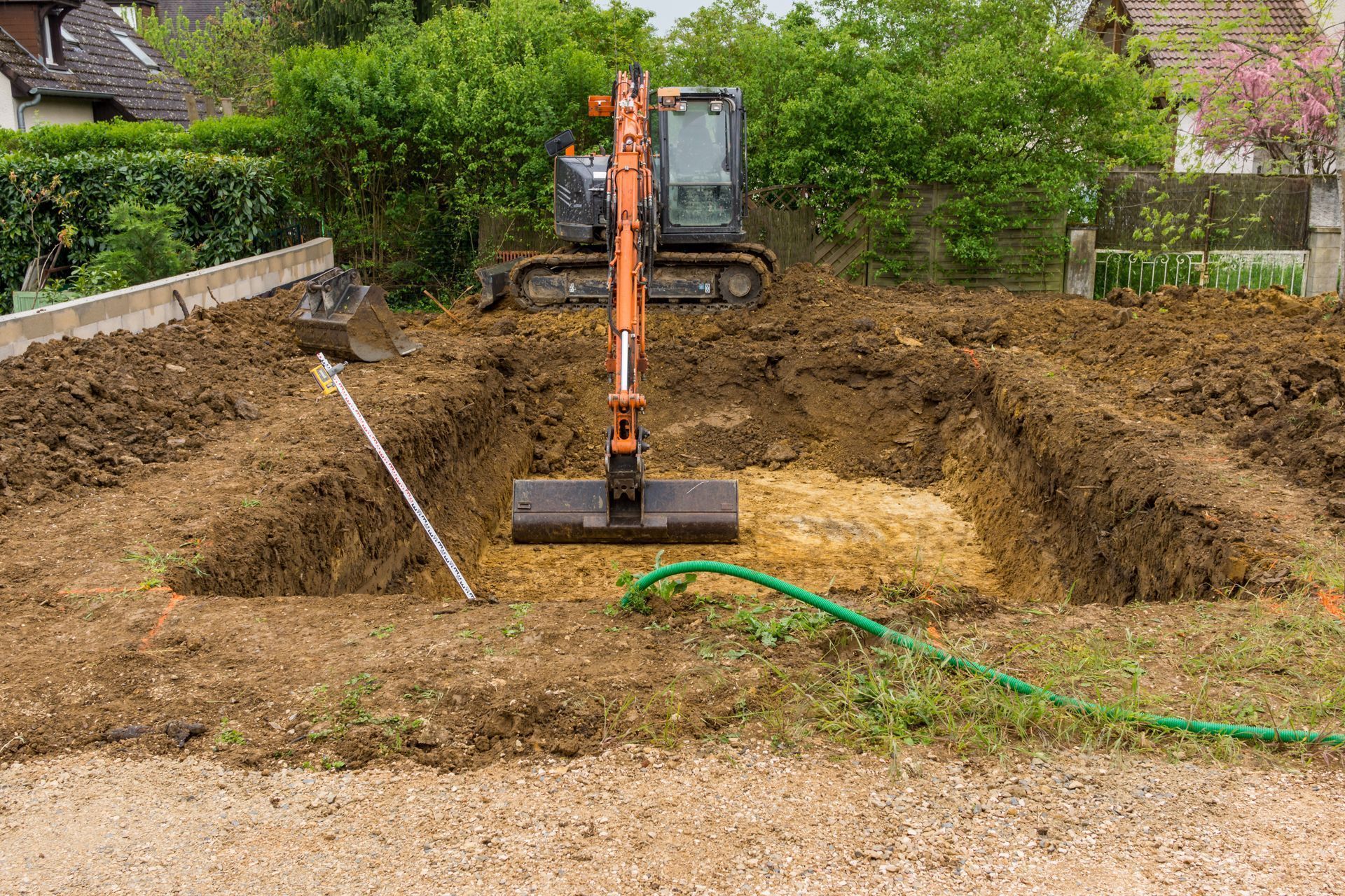 Excavatrice creusant une fondation rectangulaire dans une cour, avec des tas de terre et un tuyau à proximité.