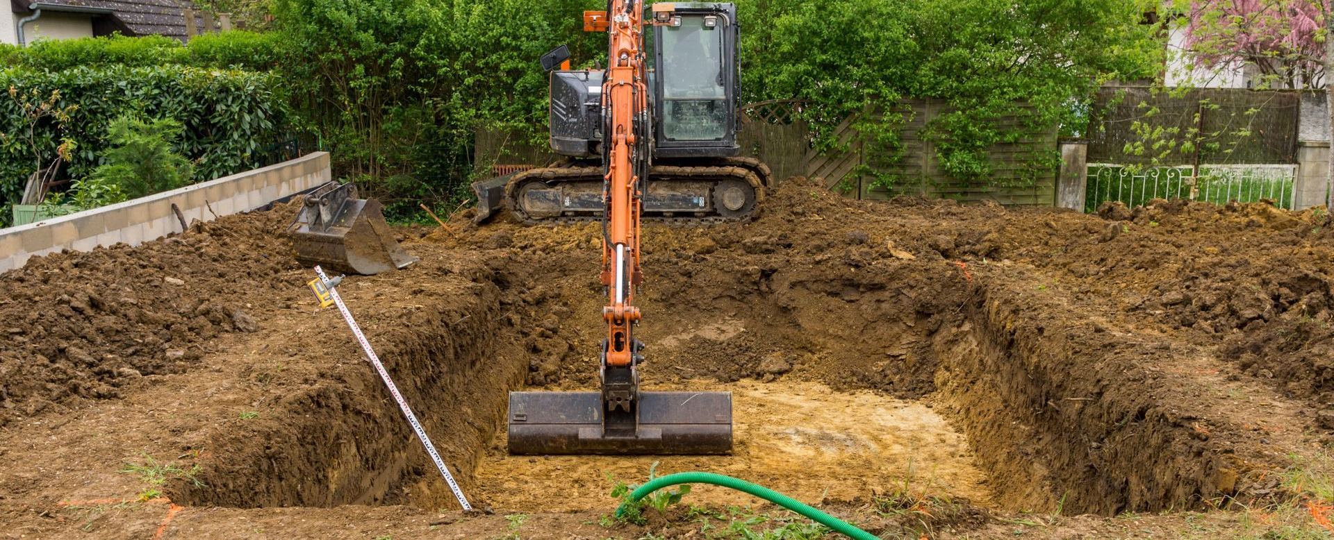 Une excavatrice creuse un trou rectangulaire dans la terre. Un tuyau vert et une bordure en béton sont visibles dans le décor ensoleillé.