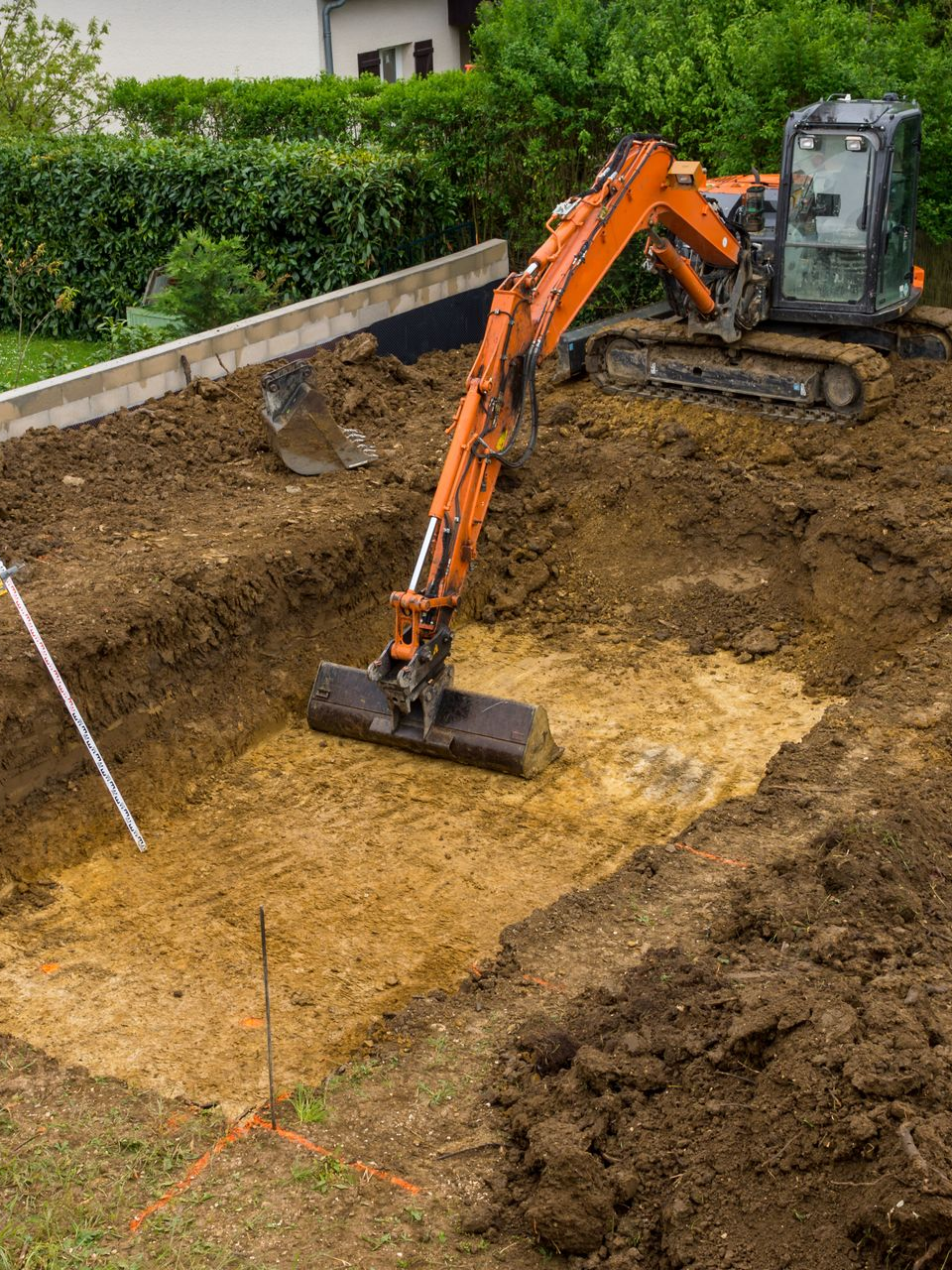 Une excavatrice orange creuse une fosse rectangulaire dans une cour. Un feuillage vert et une maison sont visibles en arrière-plan.