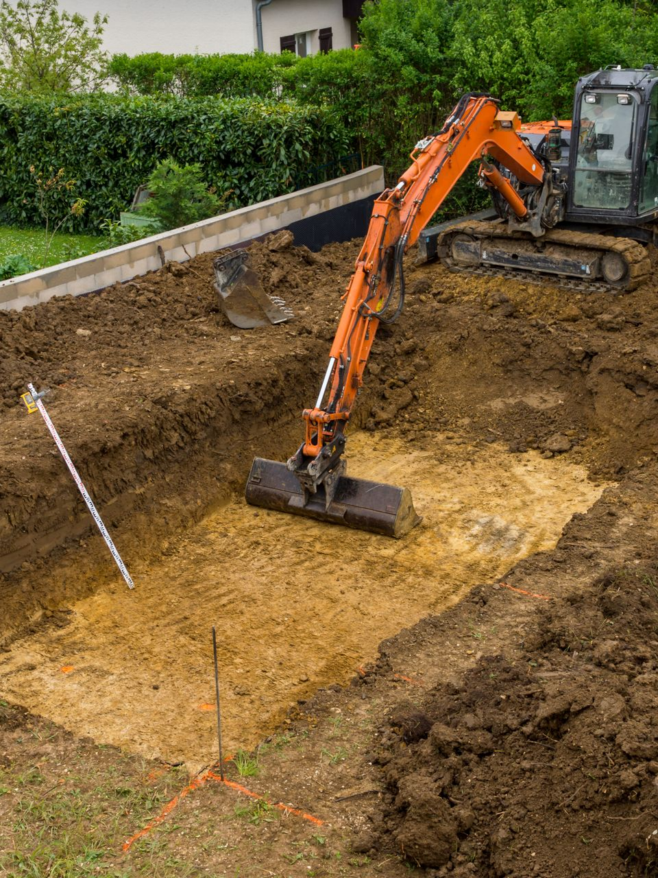 Une excavatrice orange creuse une fosse rectangulaire. Une barre niveleuse est posée au milieu de la terre et de l'herbe verte.