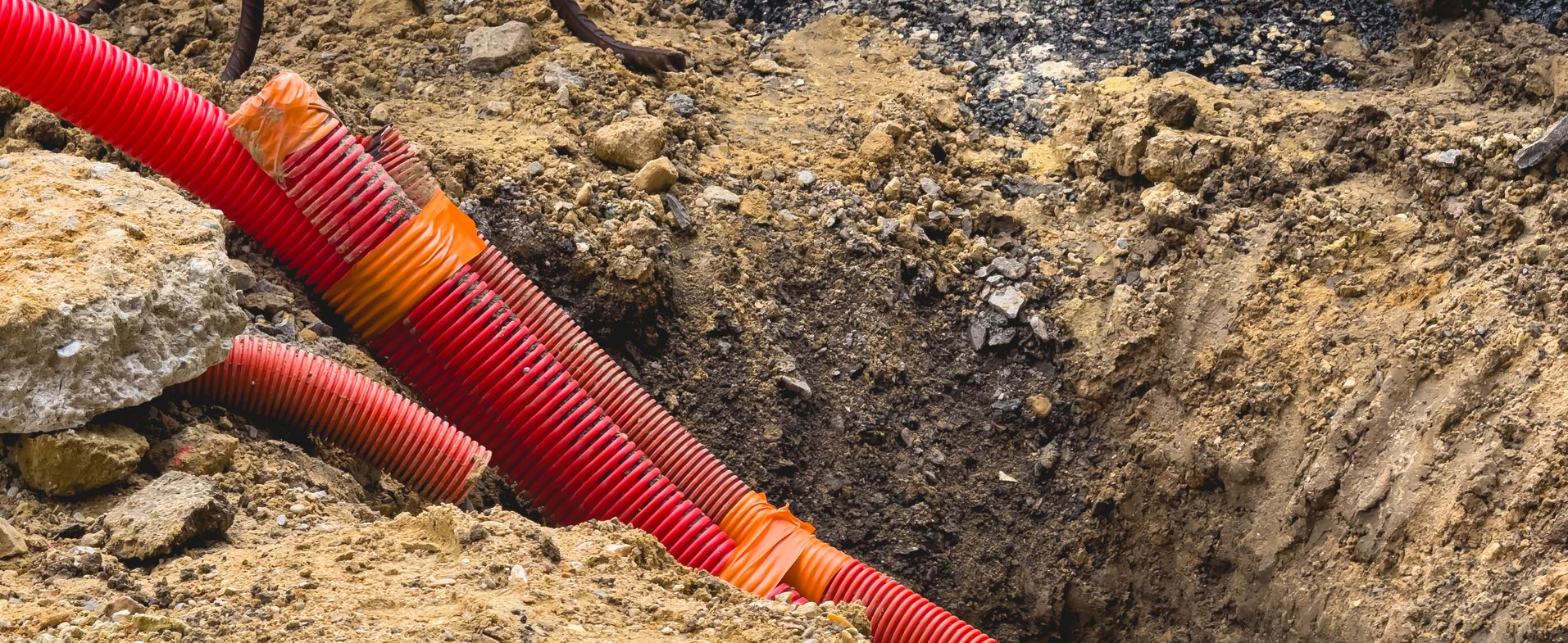 Tuyaux ondulés rouges dans une tranchée ouverte, entourés de terre et de petits rochers.