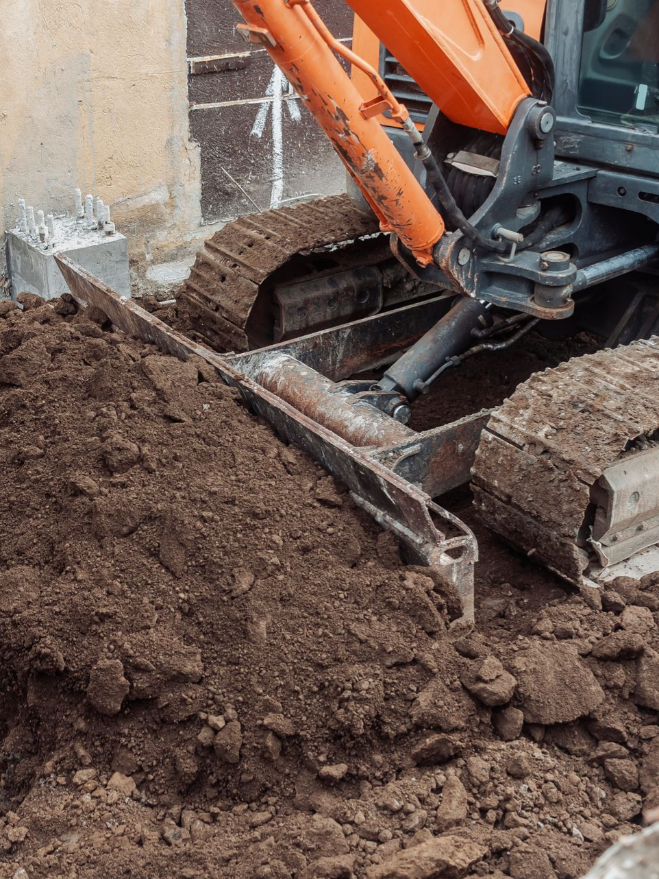 Une excavatrice orange creuse dans un tas de terre brun foncé près des fondations d'un bâtiment.
