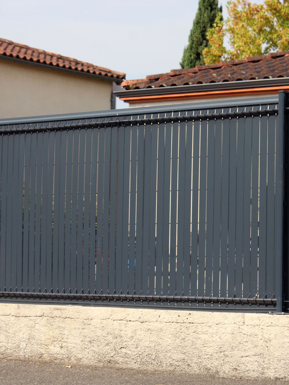 Portail coulissant en métal gris à lames verticales, devant une maison.