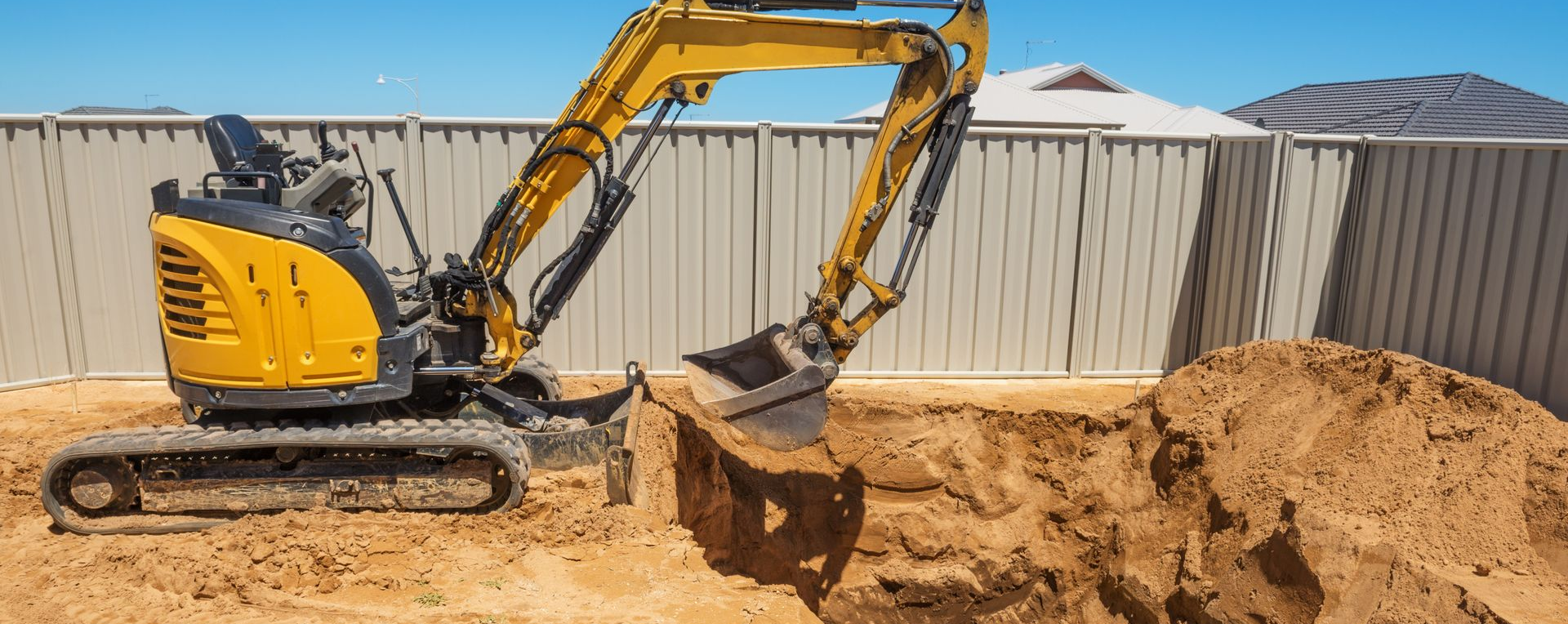 Une excavatrice jaune creuse une tranchée près d'une clôture. Journée ensoleillée.