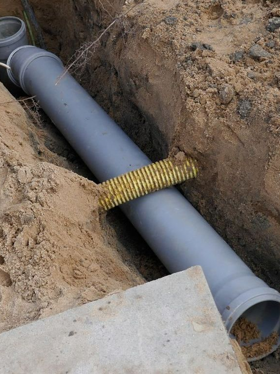 Tranchée étroite creusée dans la terre, contenant des tuyaux gris, près d'une route et d'un trottoir.
