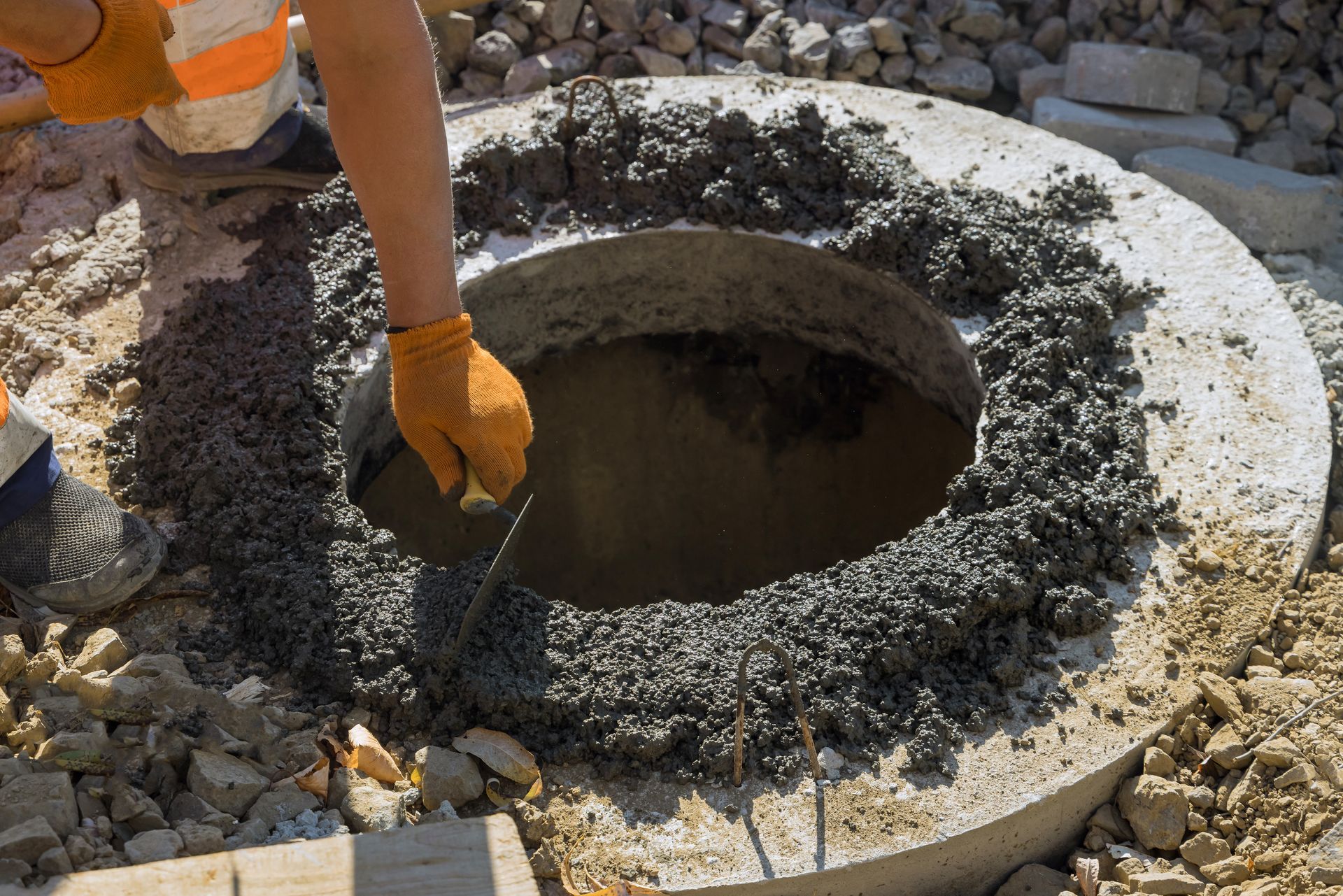 Une personne portant des gants orange applique du ciment autour d'un couvercle de regard en béton sur un chantier de construction.