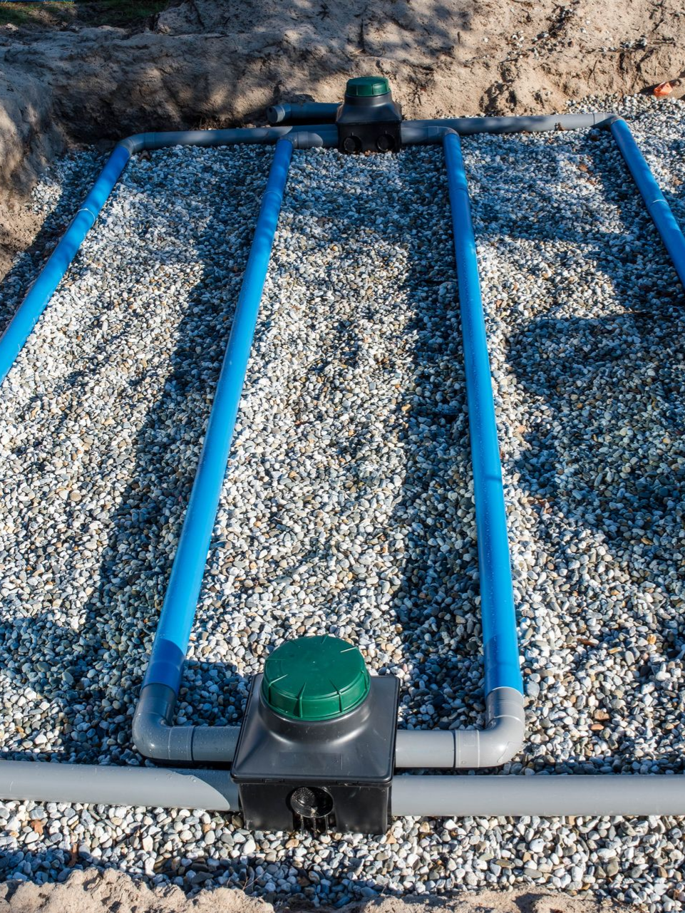 Tranchée remplie de gravier avec tuyaux bleus pour un champ d'épuration septique, entourée de terre, avec deux boîtes d'accès à capuchon vert.