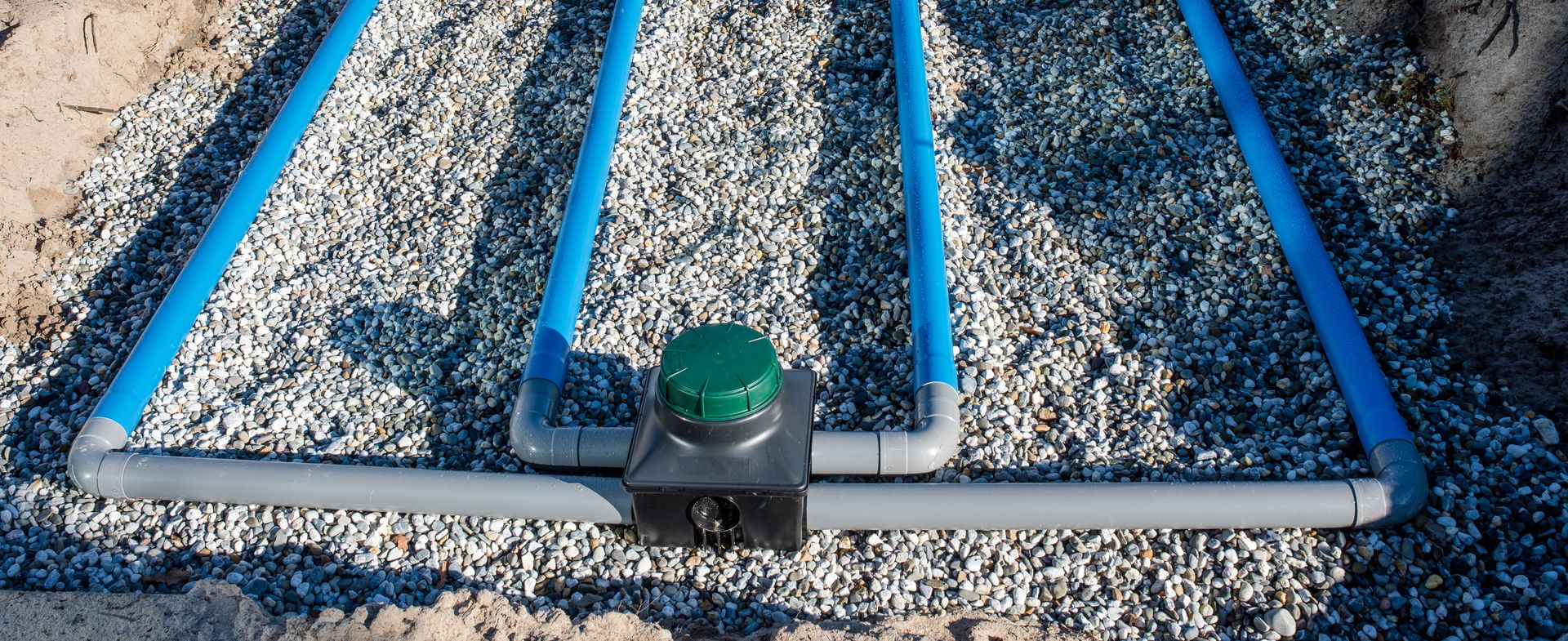 Tuyaux bleus et gris sur gravier. Une boîte à couvercle vert trône au centre.