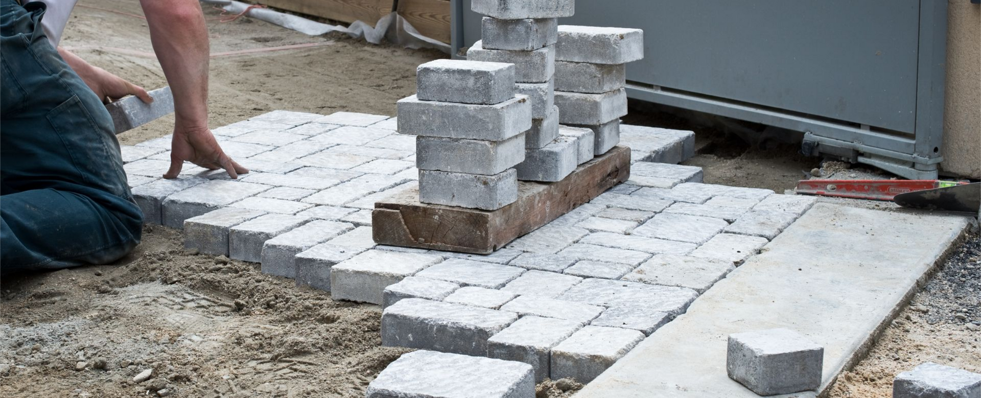 Personne posant des pavés en extérieur. Pierres grises sur du sable, avec des piles de pierres à proximité.