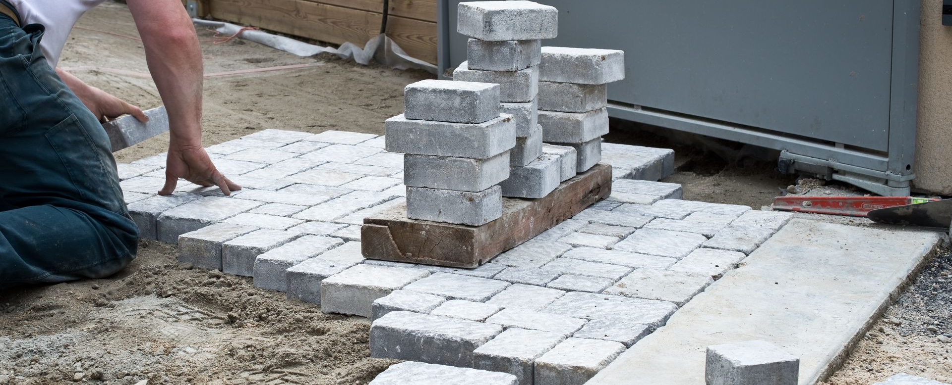 Une personne pose des pavés sur un sol sablonneux. Une pile de pierres repose sur une palette en bois.