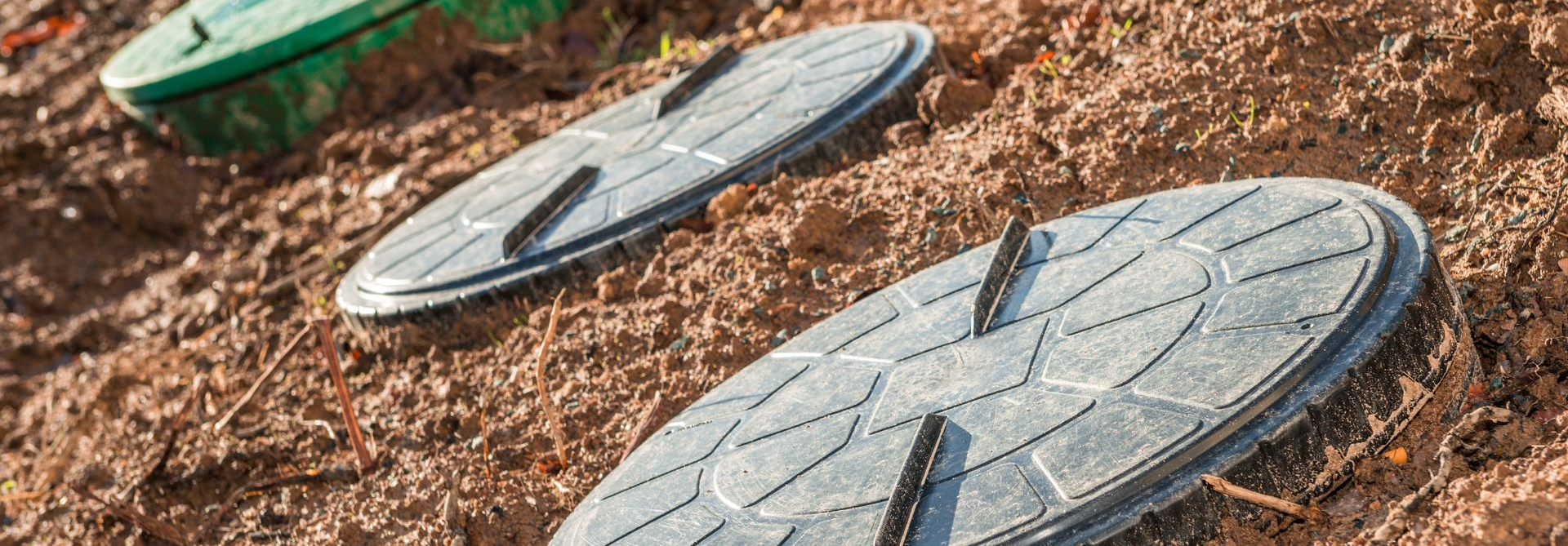 Trois plaques d'égout grises, rondes et texturées, posées sur un sol en terre brune. Une plaque d'égout verte est visible à l'arrière-plan.