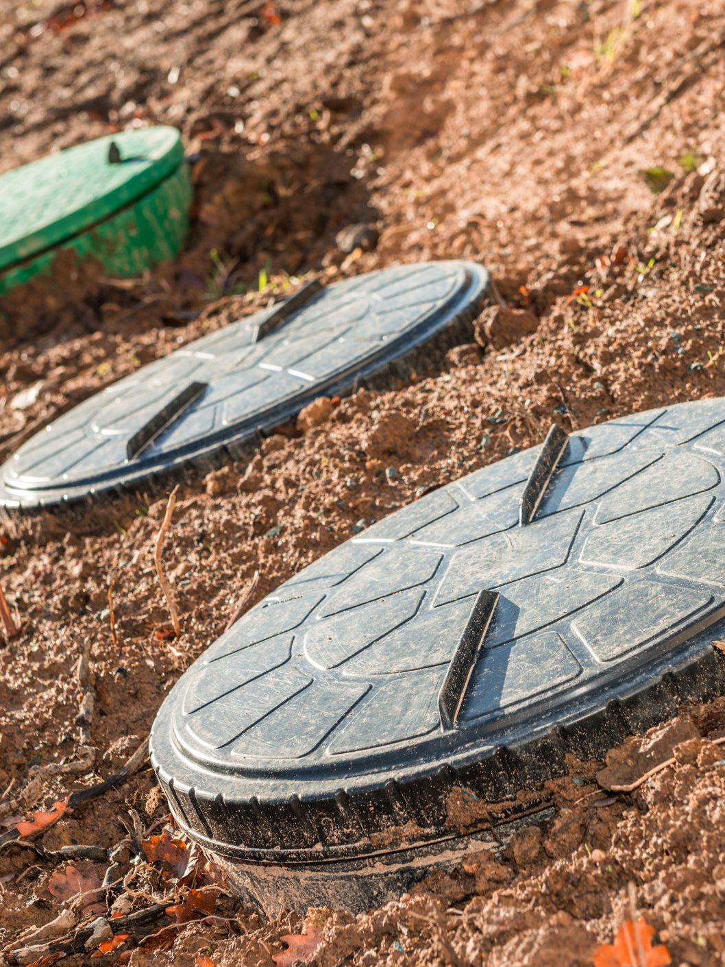 Trois couvercles de fosse à toutes eaux ovales et sombres partiellement enterrés dans la terre, l'un d'eux est vert en arrière-plan.