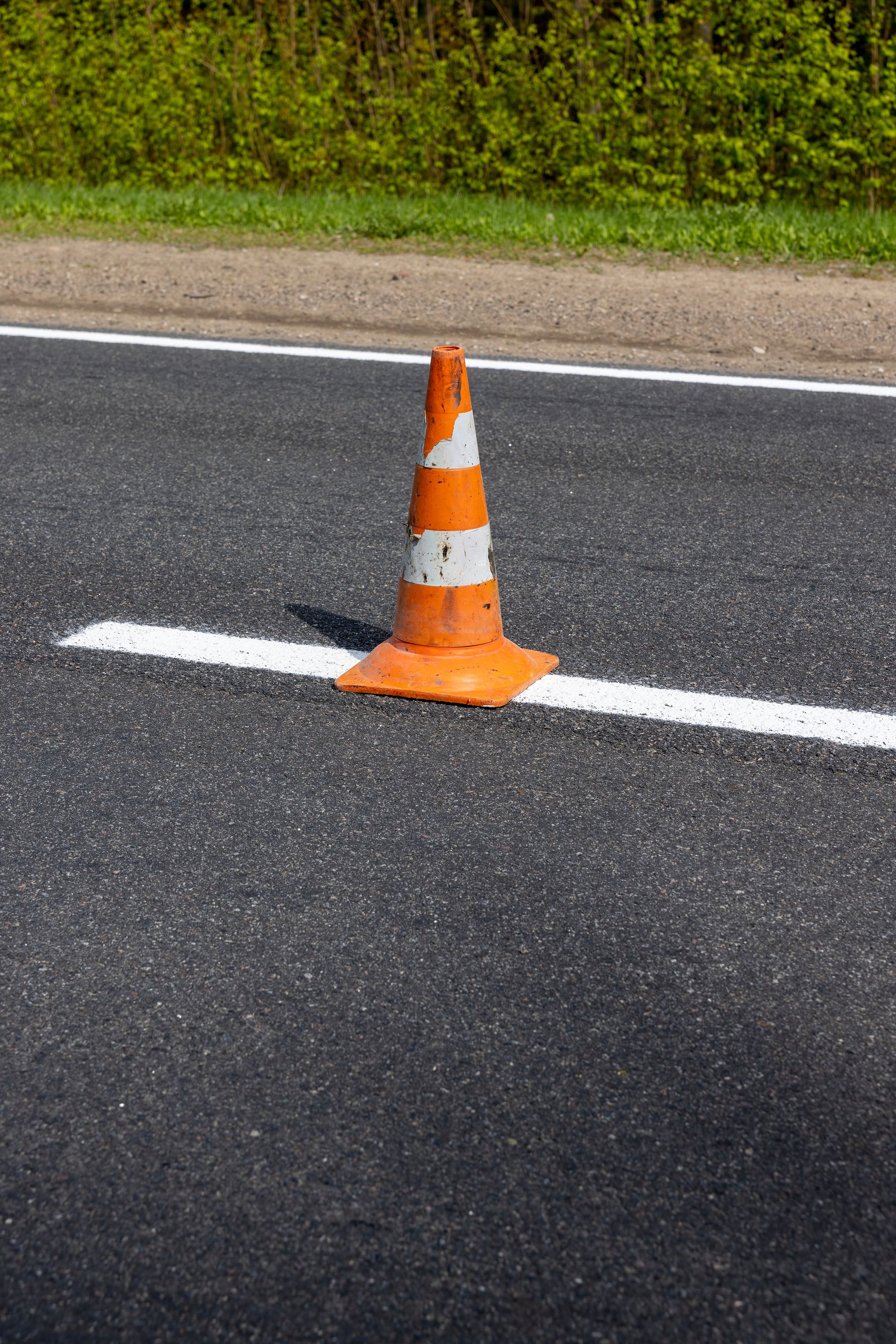 Cône de signalisation orange et blanc sur une route asphaltée nouvellement pavée, à côté d'une ligne blanche ; feuillage vert en arrière-plan.