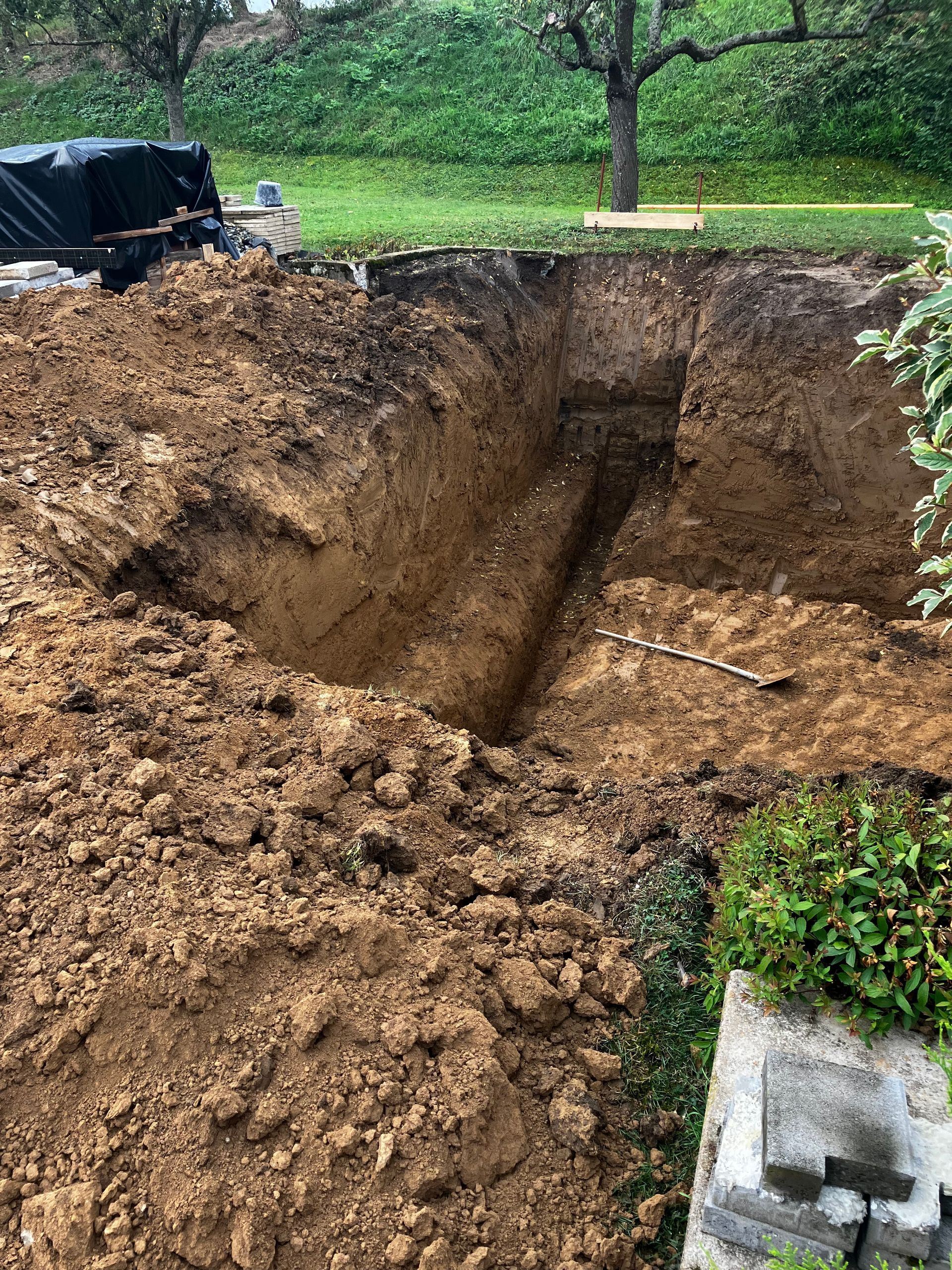 Un trou de construction ouvert creusé dans une cour avec des tas de terre et de l'herbe verte en arrière-plan.
