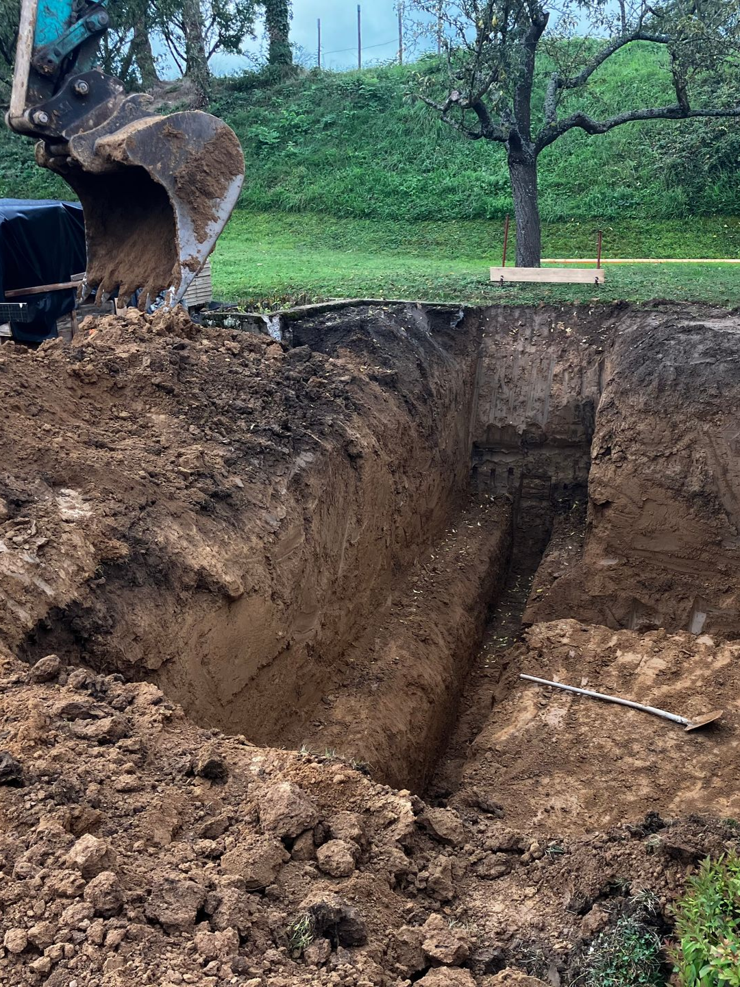 Une excavatrice creuse une tranchée profonde dans une zone herbeuse ; un tas de terre au premier plan, un arbre à l'arrière-plan.