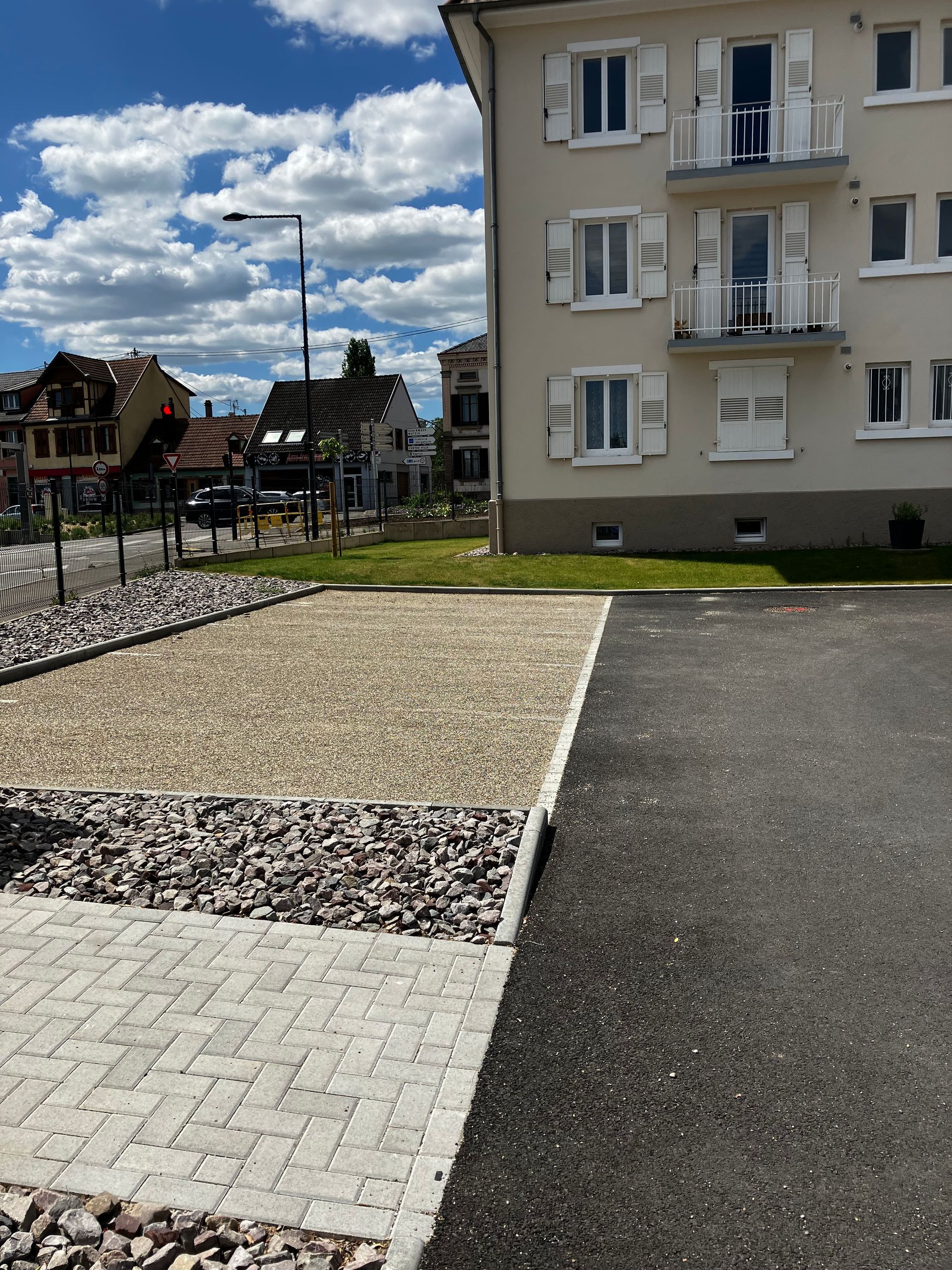Immeuble d'appartements à côté de zones de gravier, de briques et d'asphalte, sous un ciel nuageux.