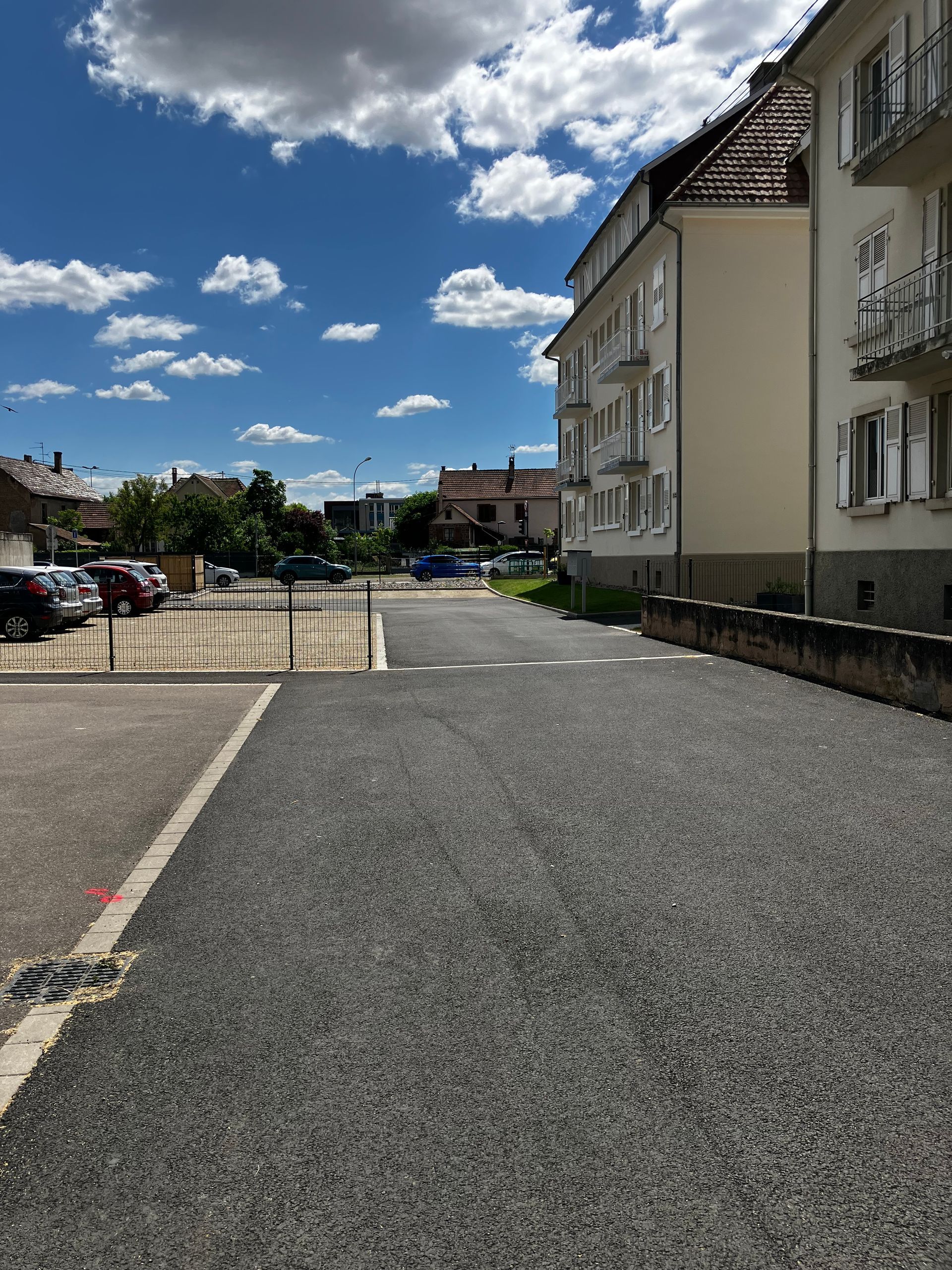 Parking en gravier à côté d'un immeuble couleur crème, par une journée ensoleillée. Voitures garées au loin.