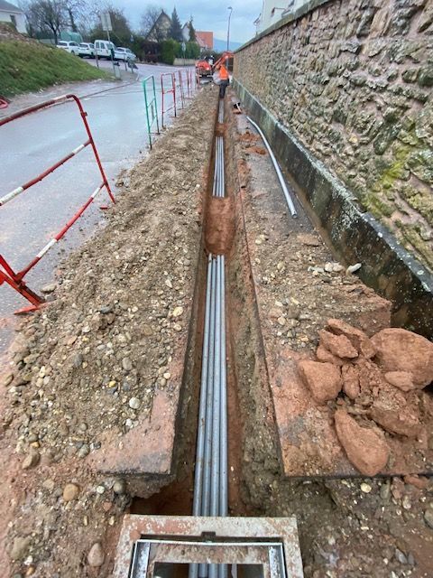 Chantier : des ouvriers installent des câbles dans une tranchée le long d'une rue, près d'un mur de pierre.