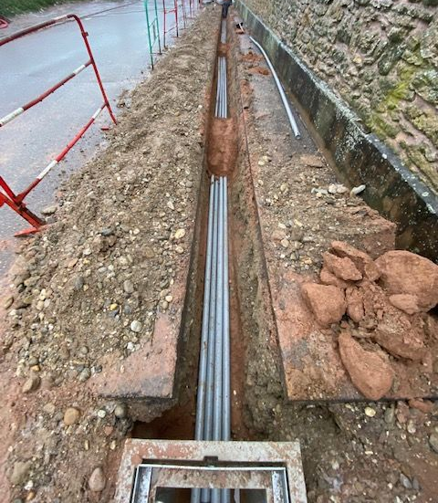 Tranchée étroite avec tuyaux gris, à côté d'une route avec barrières rouges et vertes.
