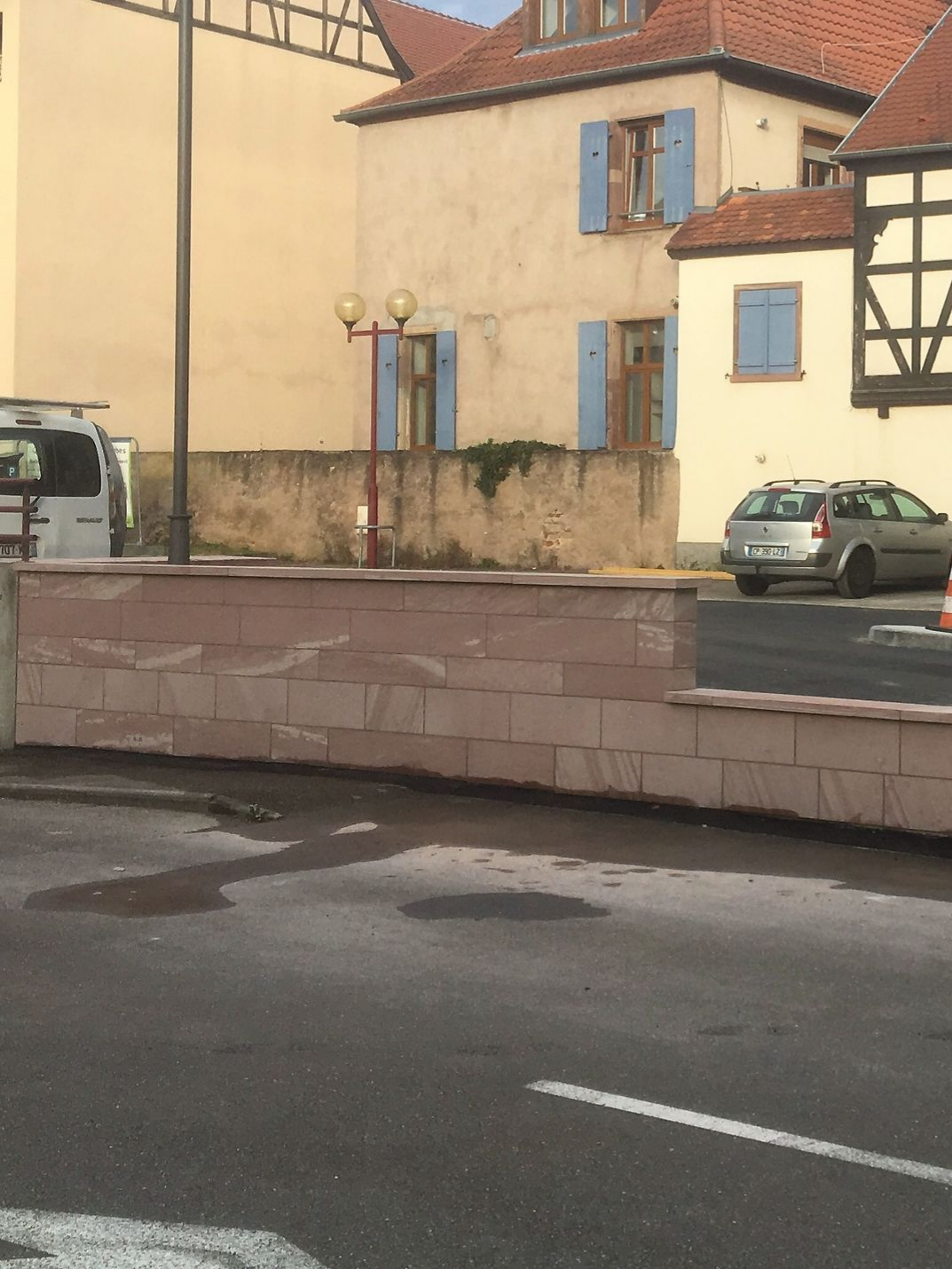Mur de pierre devant des bâtiments aux volets bleus ; voiture garée dans la rue.