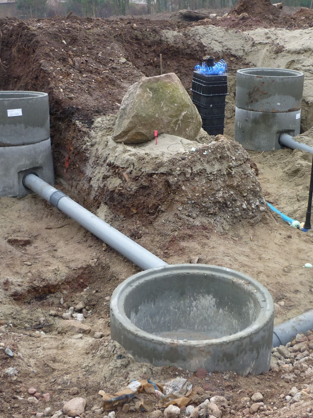 Chantier de construction avec anneaux en béton, tuyaux et terre. Un gros rocher repose sur le sol.