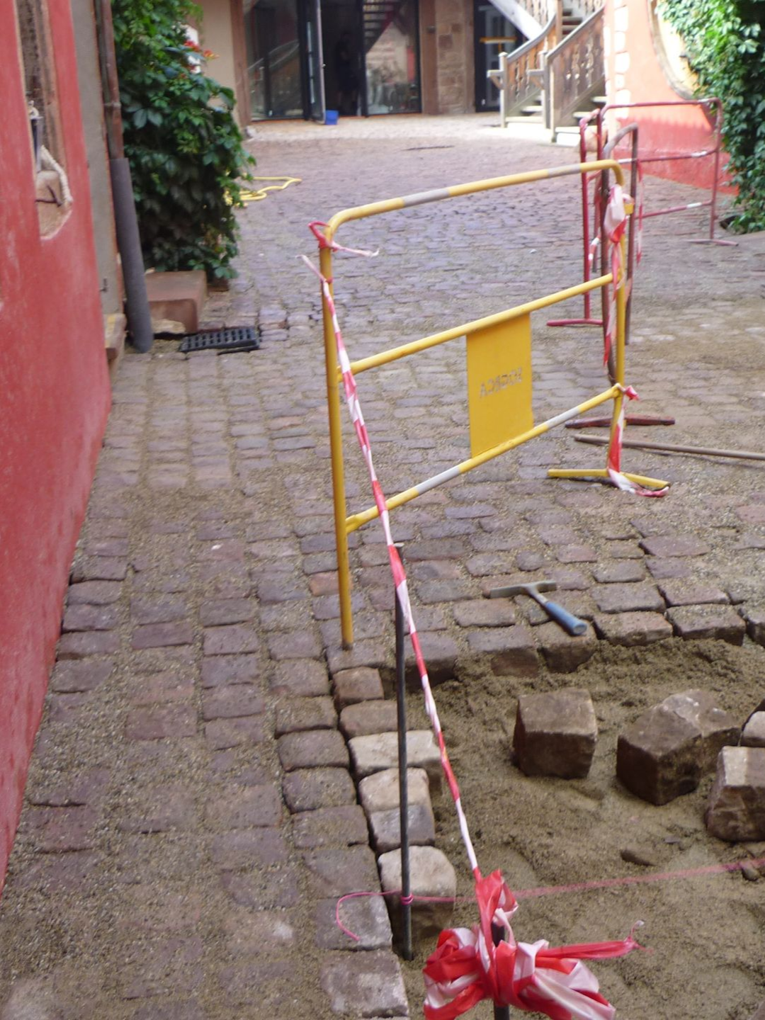 Trottoir en travaux, barré par une barrière jaune et du ruban rouge et blanc. Pavés, sable et mur de briques.