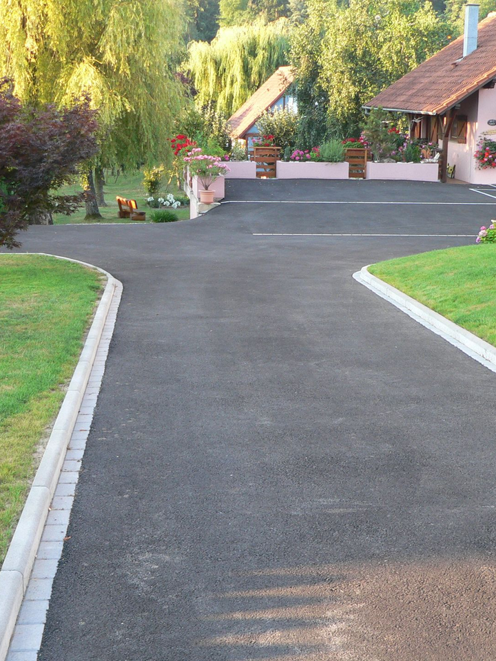 Allée asphaltée menant à une maison, bordée d'herbe et de pierres. Arbres et fleurs en arrière-plan.