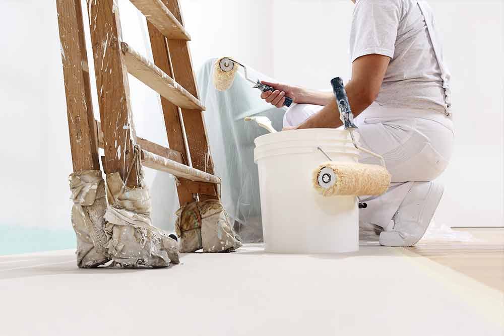 Eine Person kniet neben einem Farbeimer und einer Leiter und streicht eine Wand.