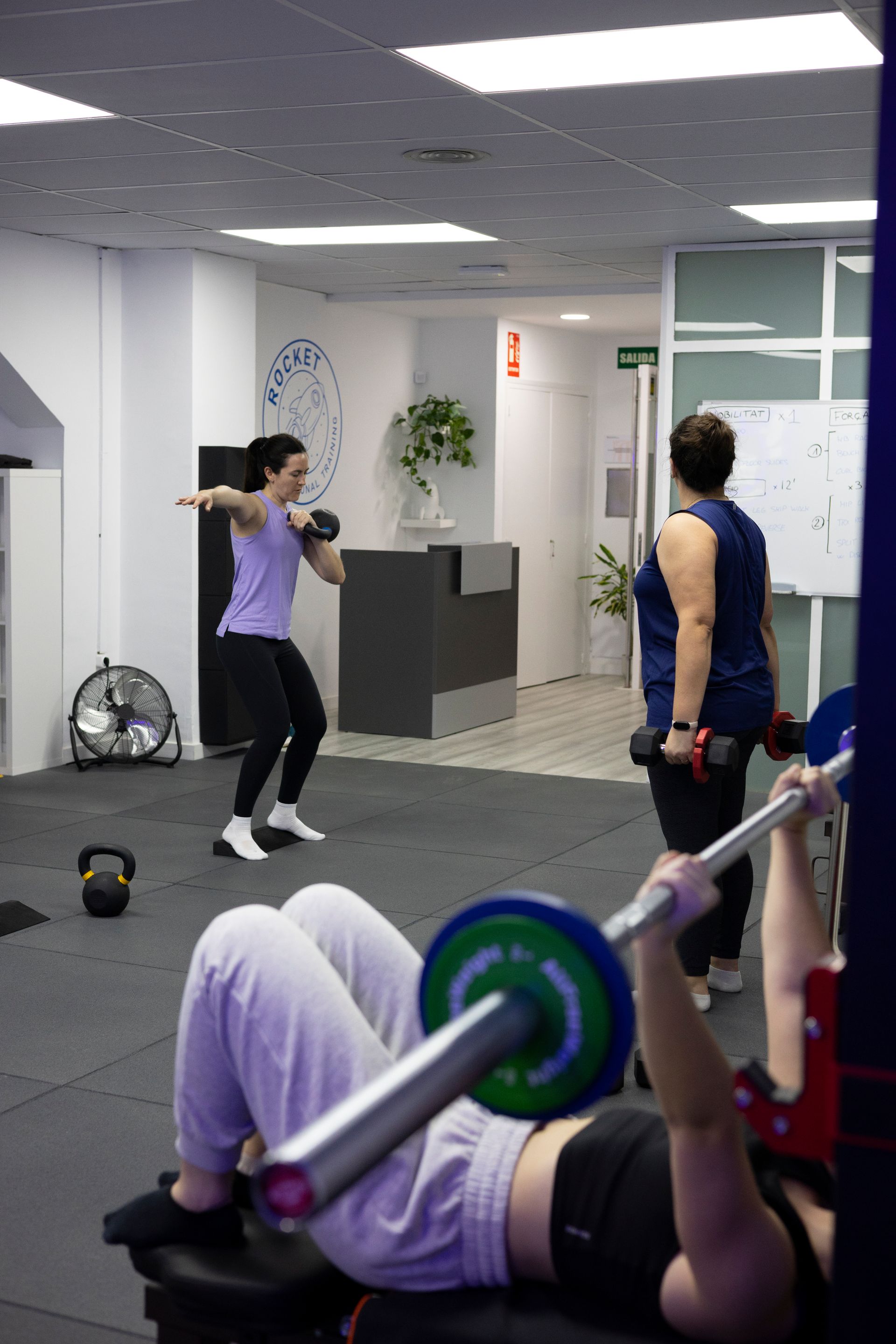 Una mujer está tumbada en un banco levantando una barra en un gimnasio.