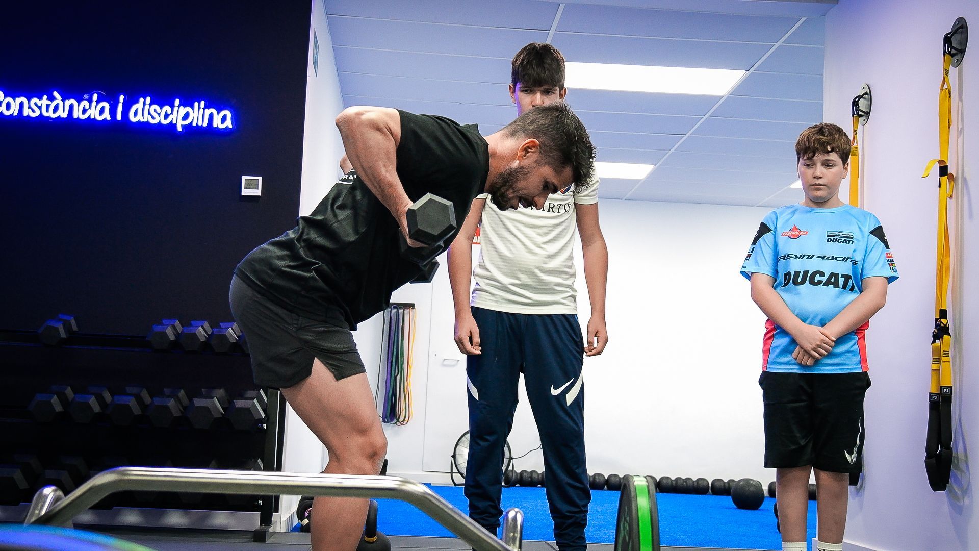 Un hombre está levantando una mancuerna en un gimnasio mientras dos niños miran.