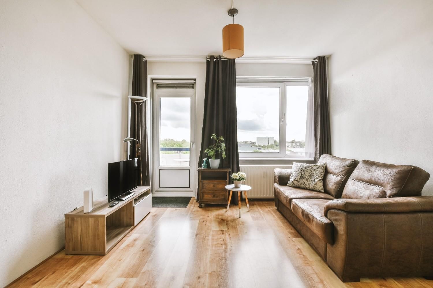 Sala de estar luminosa con sofá marrón, televisor, suelo de madera y grandes ventanales con cortinas.