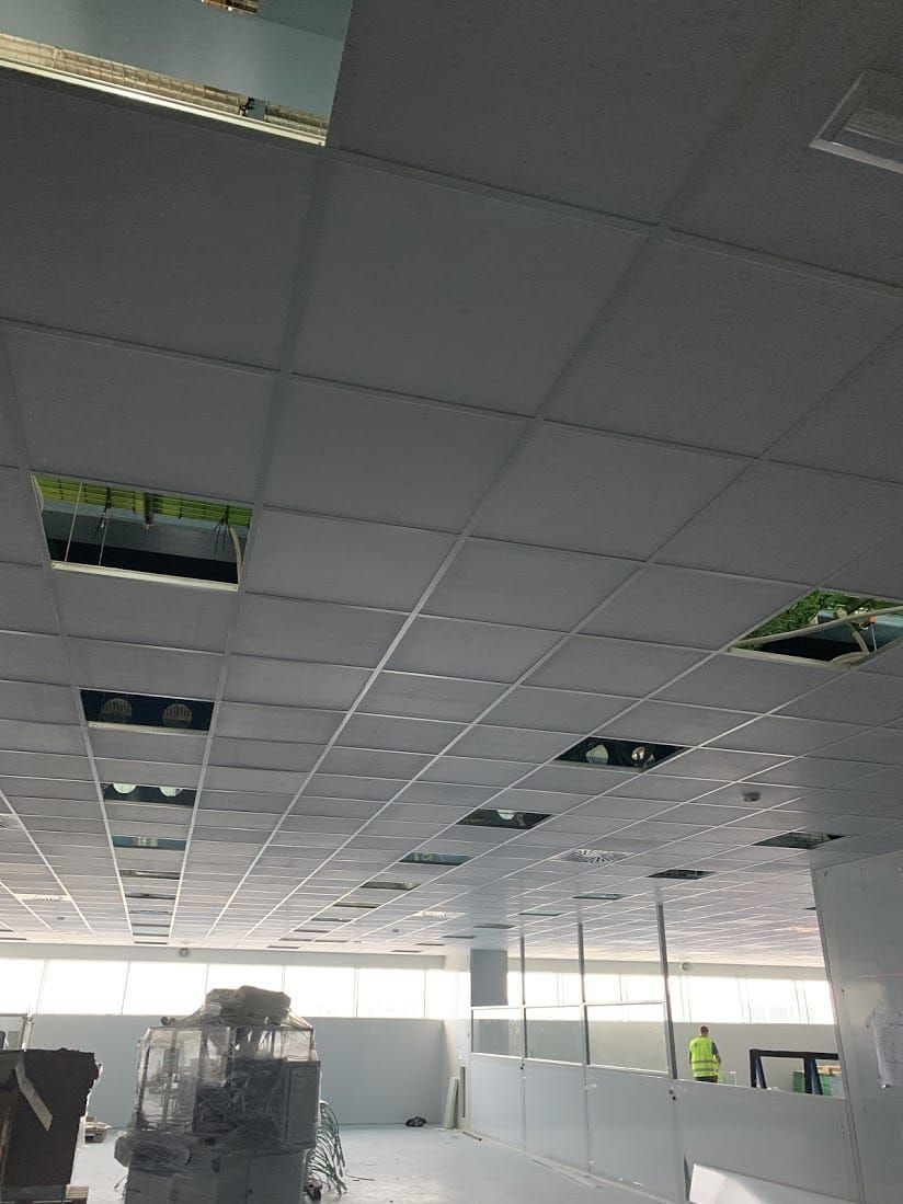 Techo de oficina dañado con baldosas faltantes y paneles expuestos, habitación luminosa debajo con trabajadores al fondo.