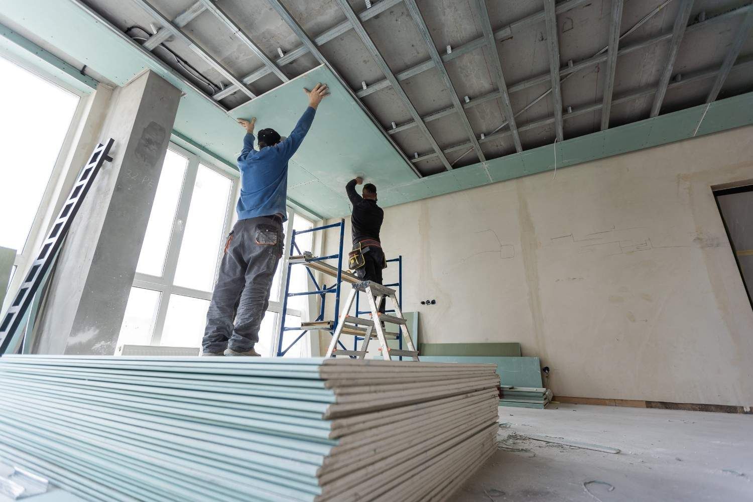 Dos trabajadores instalan paneles de yeso en el interior de una habitación sin terminar, con paneles apilados en primer plano.