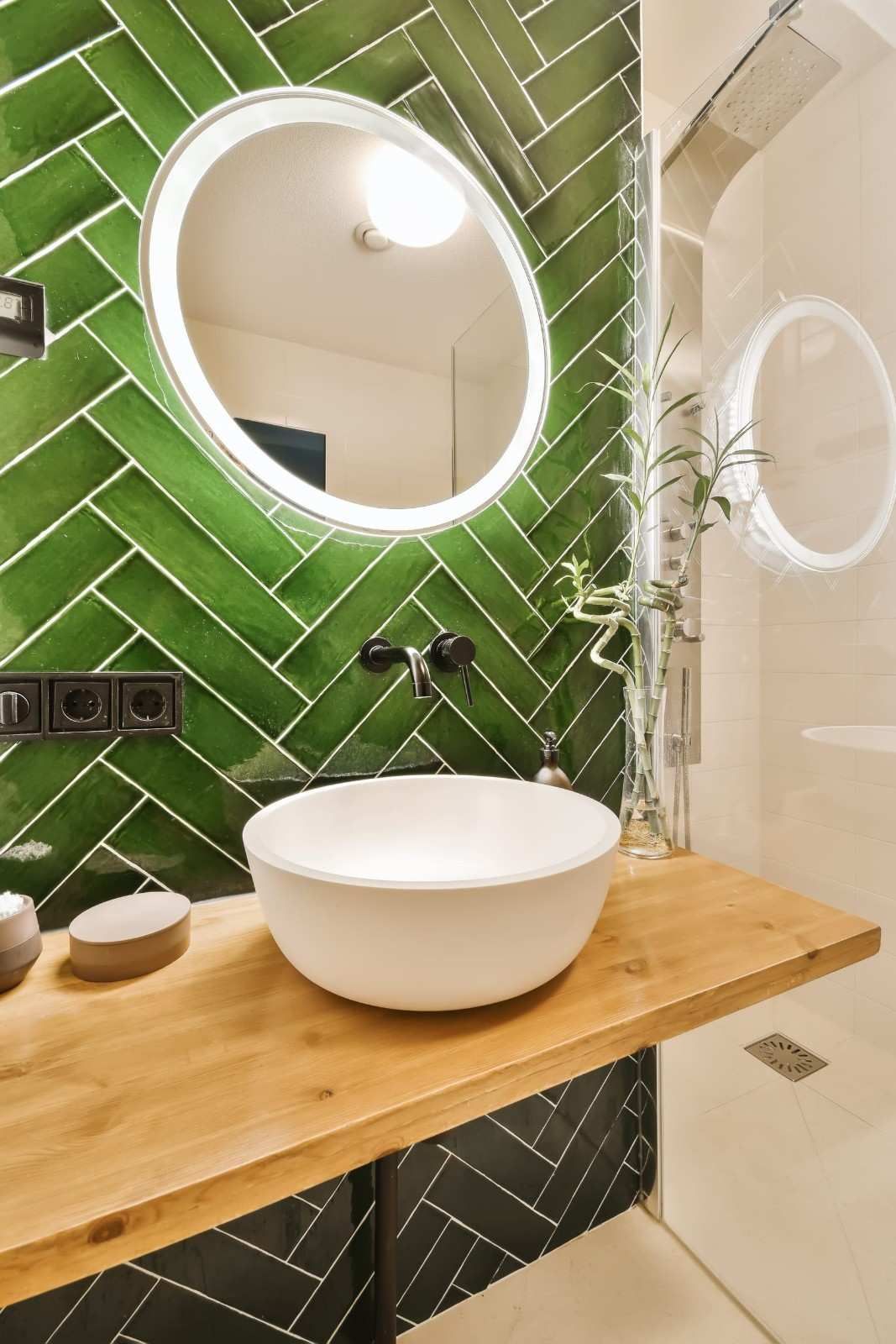 Baño moderno con pared de azulejos verdes, espejo redondo retroiluminado, lavabo blanco sobre encimera y encimera de madera.