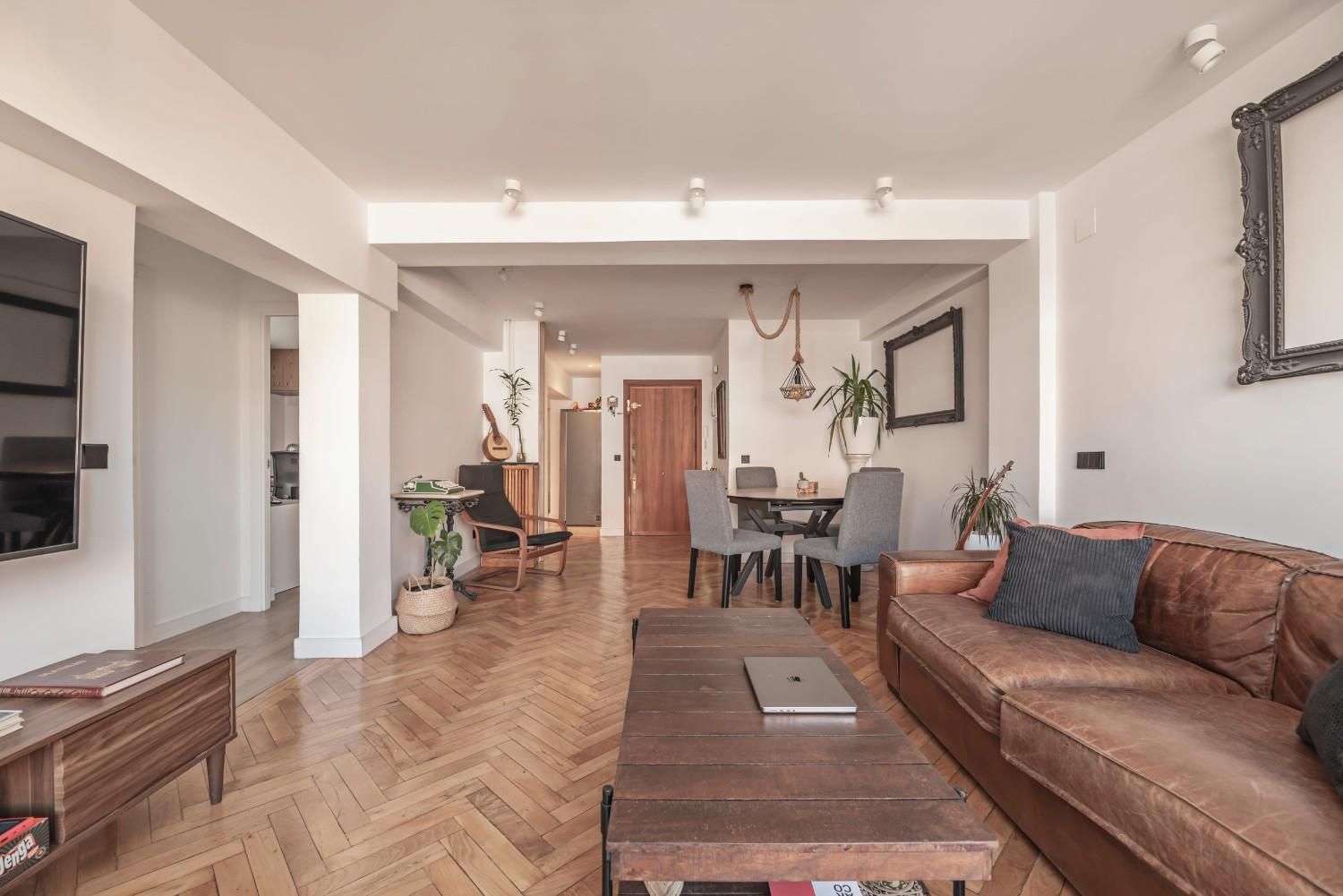 Luminoso salón con sofá de cuero marrón, mesa de centro de madera, zona de comedor y suelos de madera en espiga.