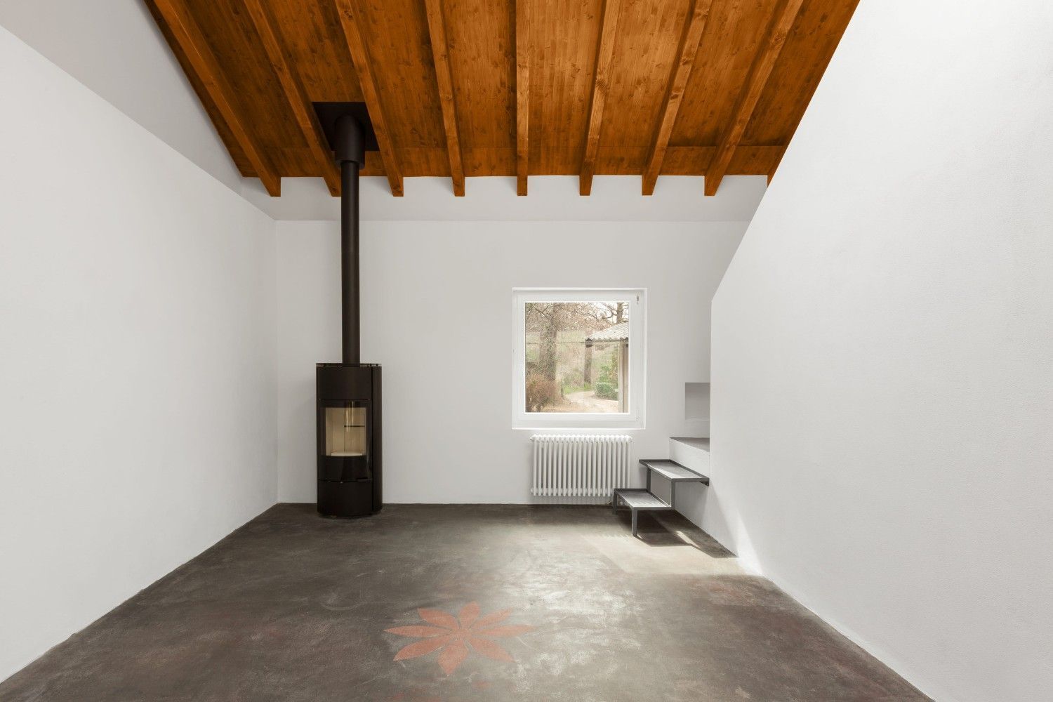 Habitación vacía con techo de madera, paredes blancas, suelo de hormigón y una pequeña estufa de leña junto a una ventana.