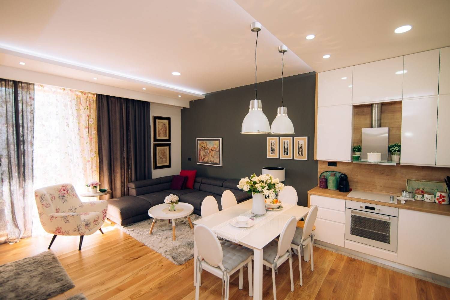 Luminoso salón-comedor moderno con muebles blancos, sofá negro, lámparas colgantes y cocina abierta.