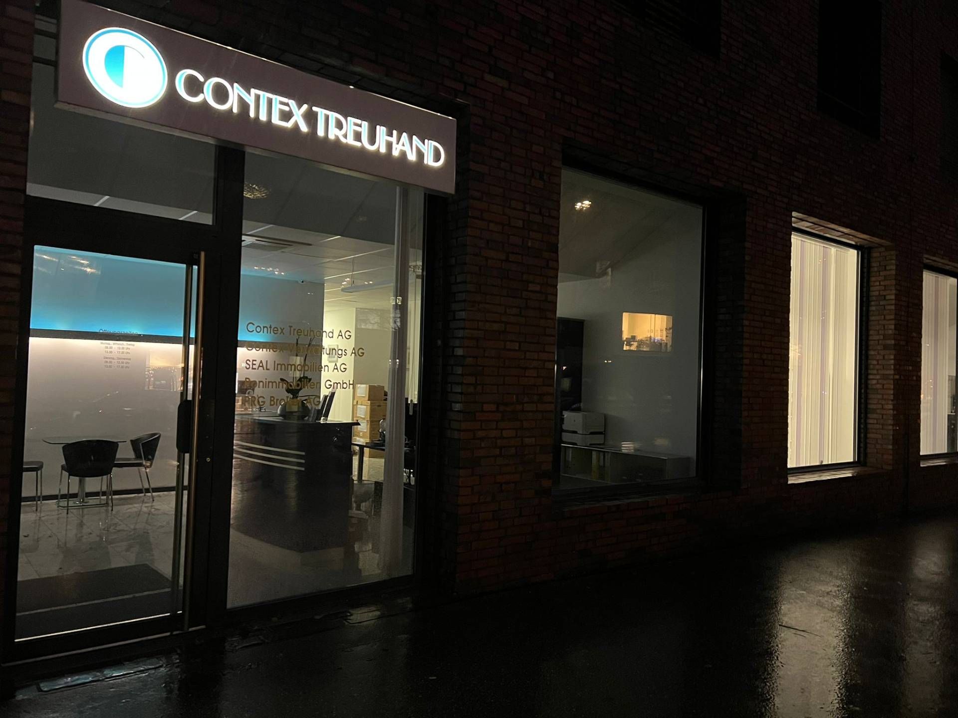 Außenansicht des Bürogebäudes „Contex Treuhand“ bei Nacht, leuchtendes Schild über den Glasfenstern. Backsteingebäude.