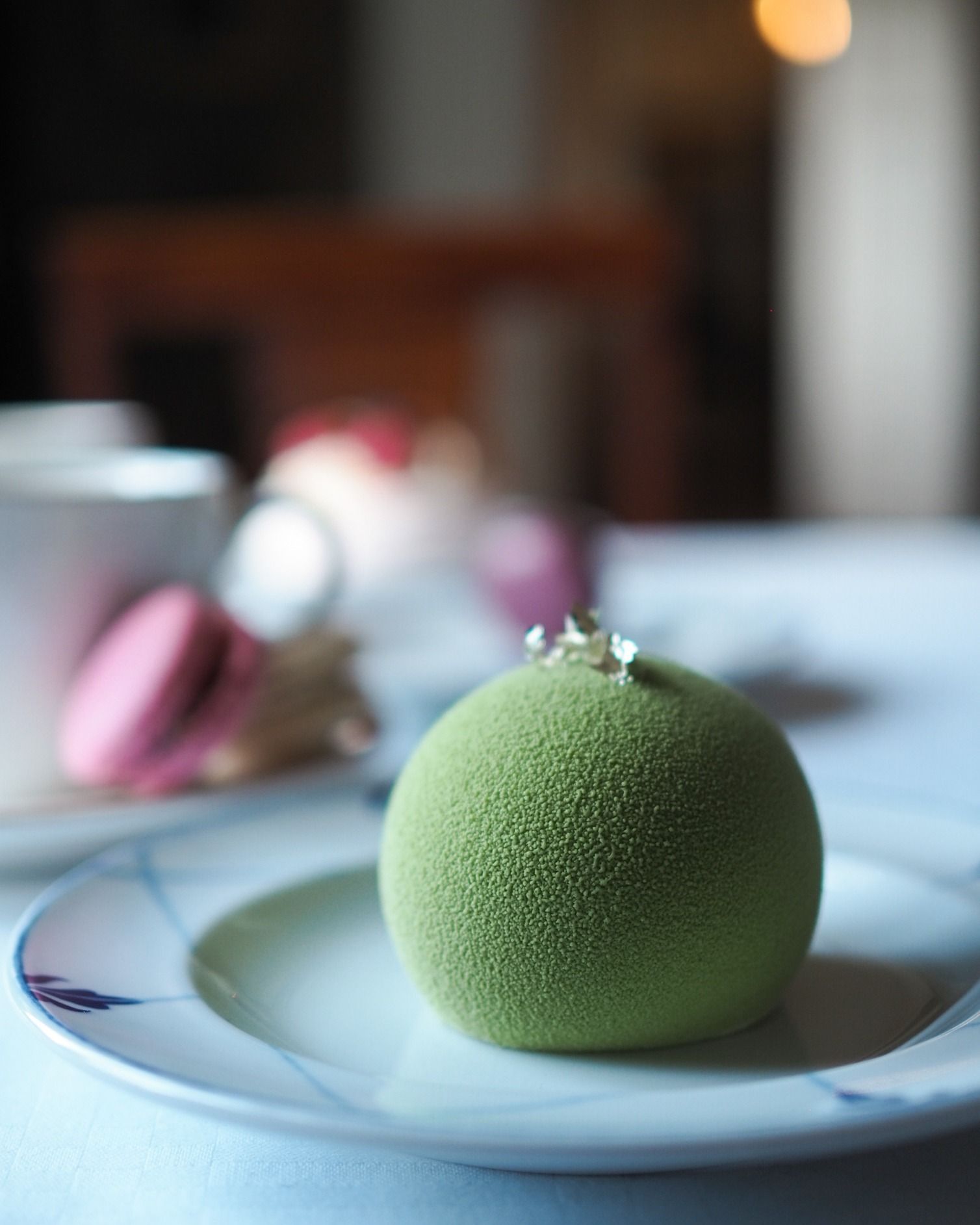 Vihreä pyöreä jälkiruoka valkoisella lautasella, taustalla sumea vaaleanpunainen macaron