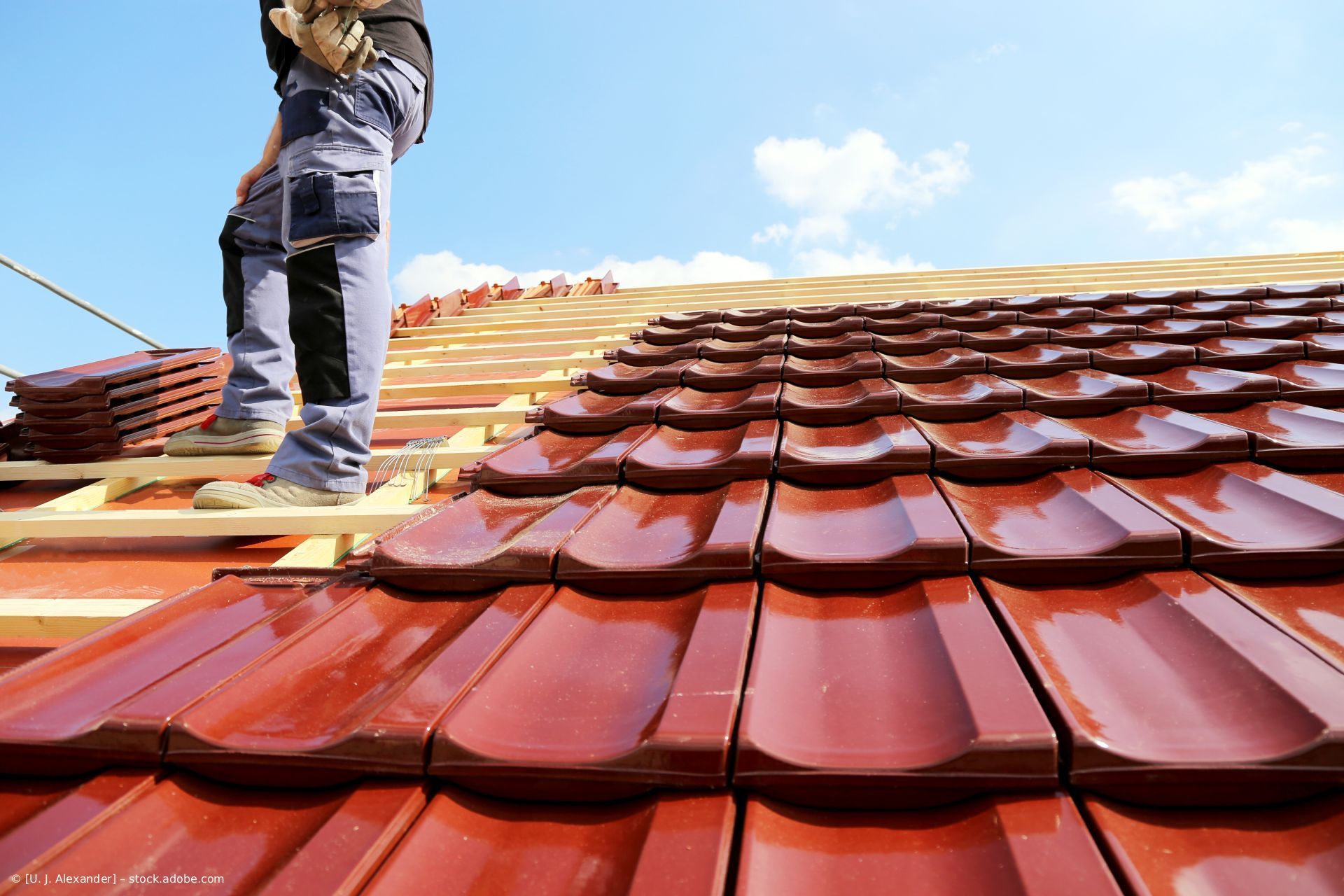 Ein Arbeiter steht auf einem im Bau befindlichen roten Ziegeldach vor blauem Himmel