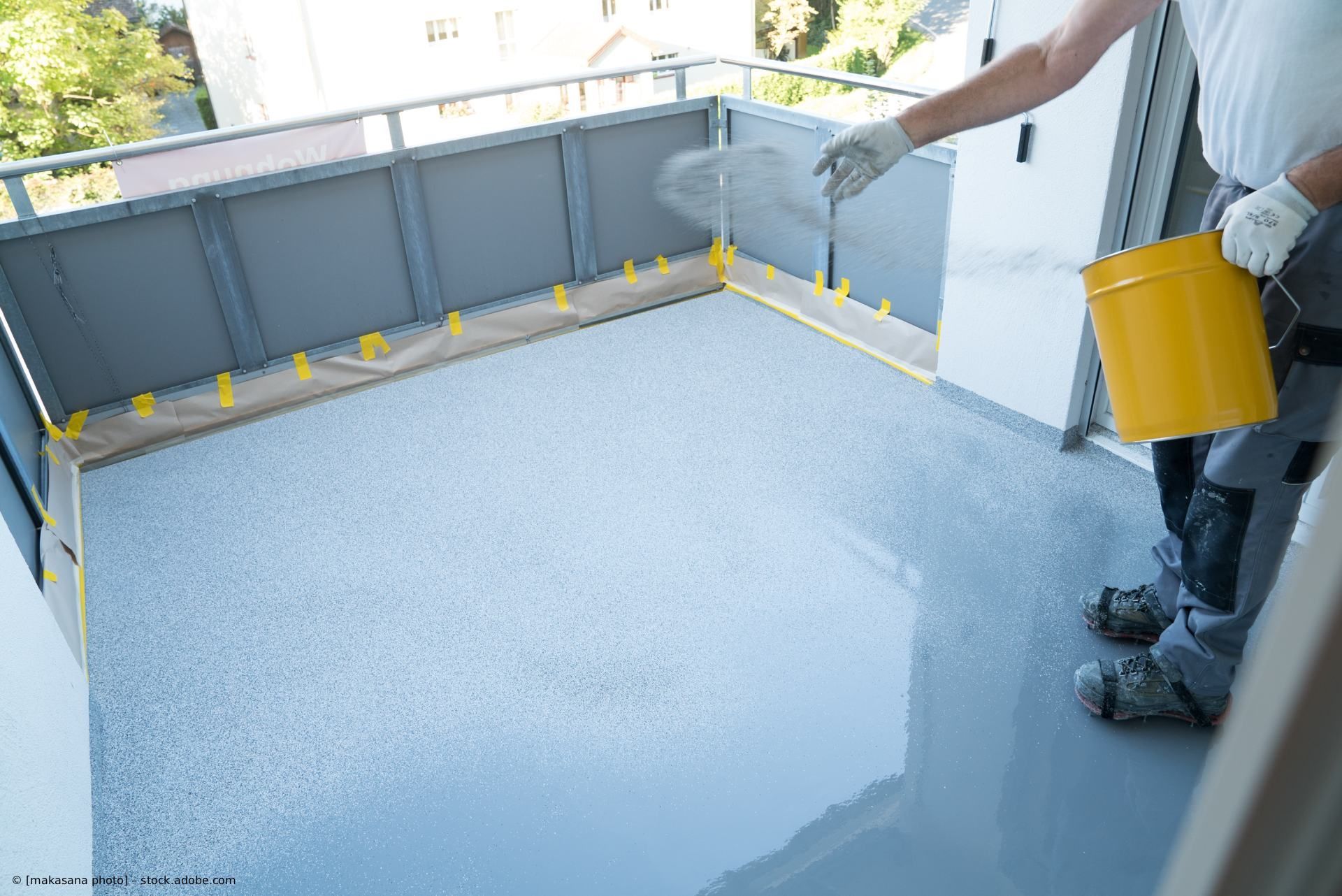Ein Arbeiter trägt auf einem Balkon eine glatte, graue Bodenbeschichtung auf und hält dabei einen gelben Eimer.