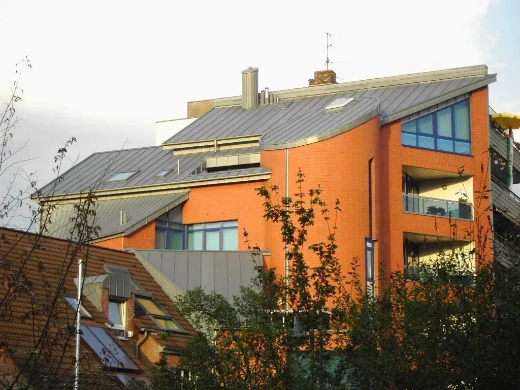 Modernes orangefarbenes Wohnhaus mit grauem Dach und blauen Fenstern, teilweise von Bäumen verdeckt.