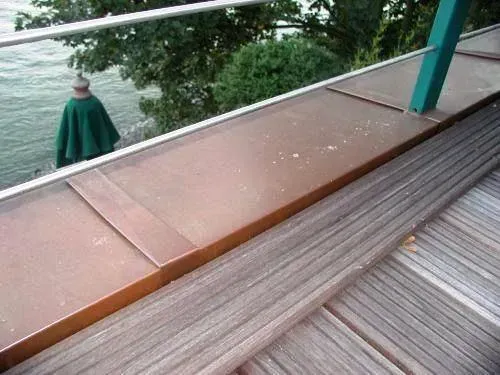 Holzterrasse und Geländer neben einem Gewässer, darunter eine Person in einem grünen Umhang.