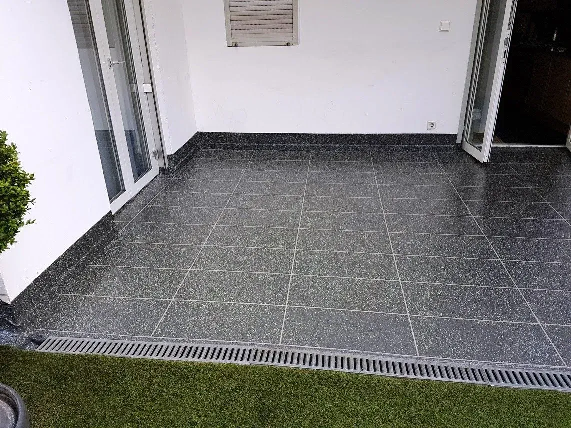 Leere, geflieste Terrasse mit Abflussrost und weißen Wänden zwischen zwei Metallpfosten