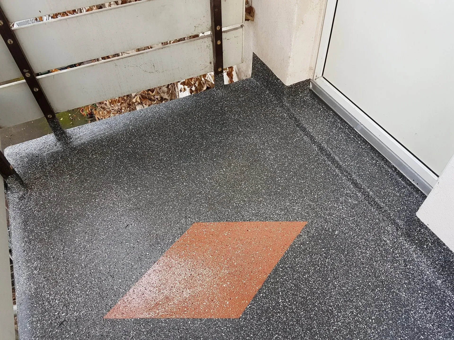 Kleine Außenecke mit grau gesprenkeltem Boden, einer orangefarbenen quadratischen Fliese und einer weißen Tür.