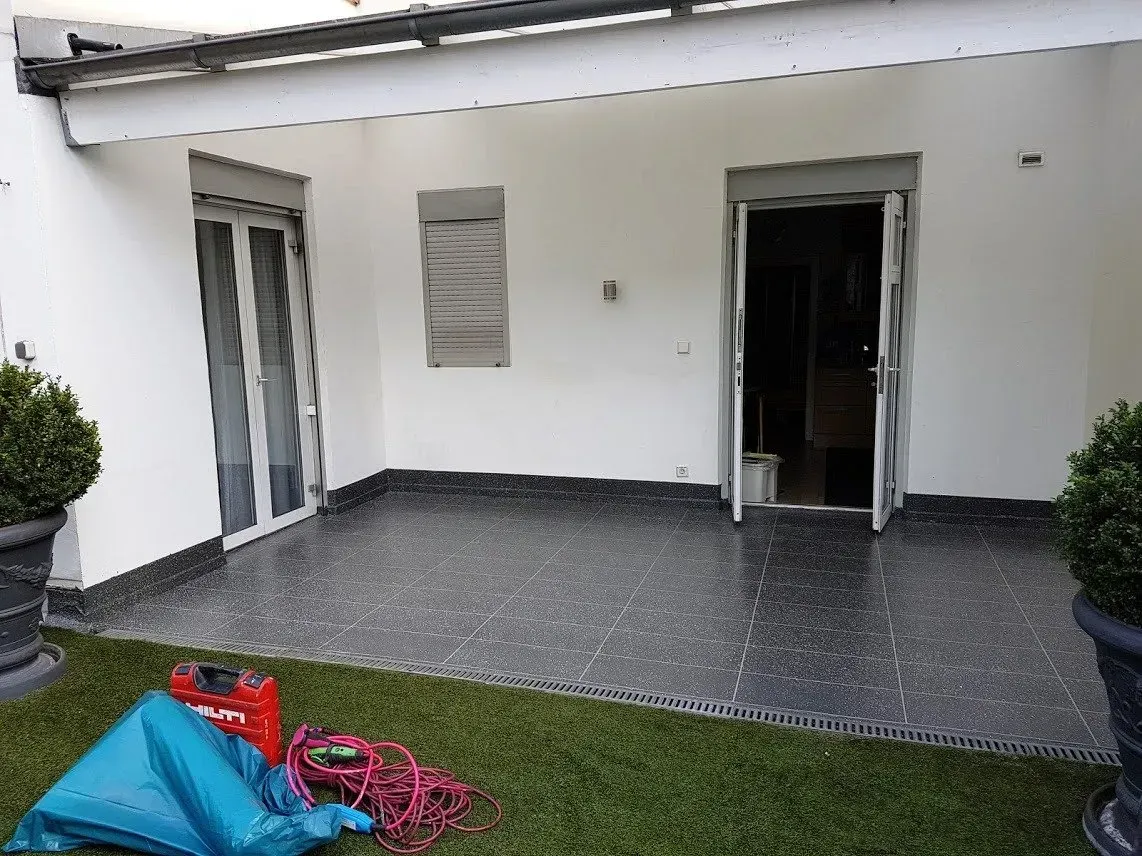 Terrasse mit grauem Fliesenboden, Glasschiebetüren und offenem Durchgang neben Topfpflanzen.