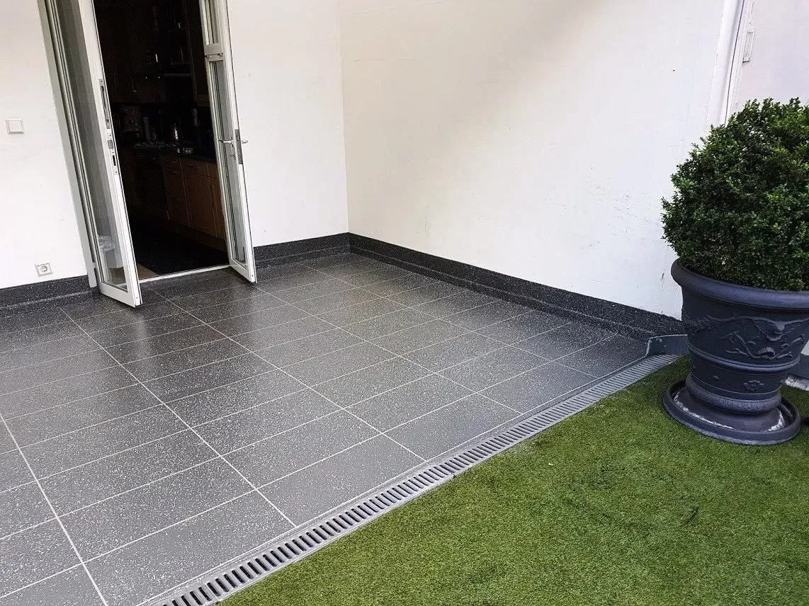 Überdachte Terrasse mit grauem Fliesenboden, weißen Wänden und einem Topfstrauch neben einem Rasenstreifen