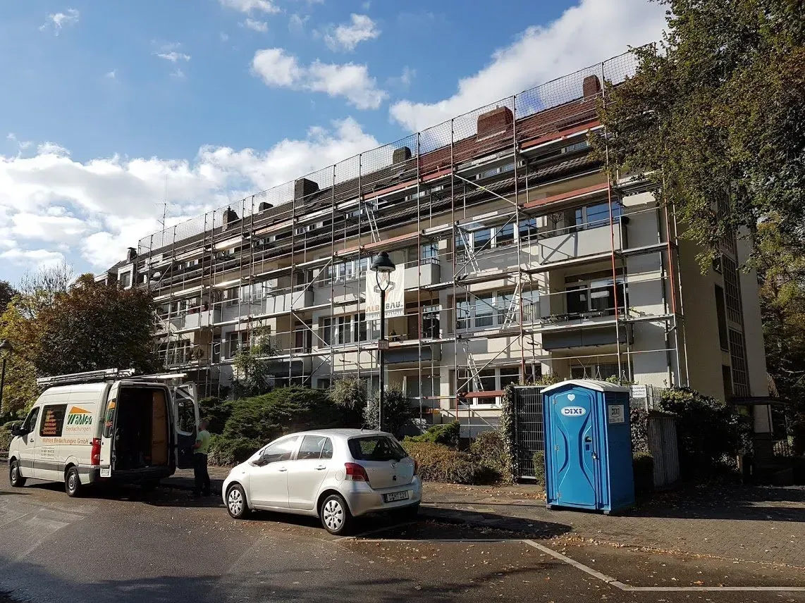 Ein Wohnhaus wird renoviert; Gerüste, geparkte Autos und eine blaue mobile Toilette stehen an einer sonnigen Straße.