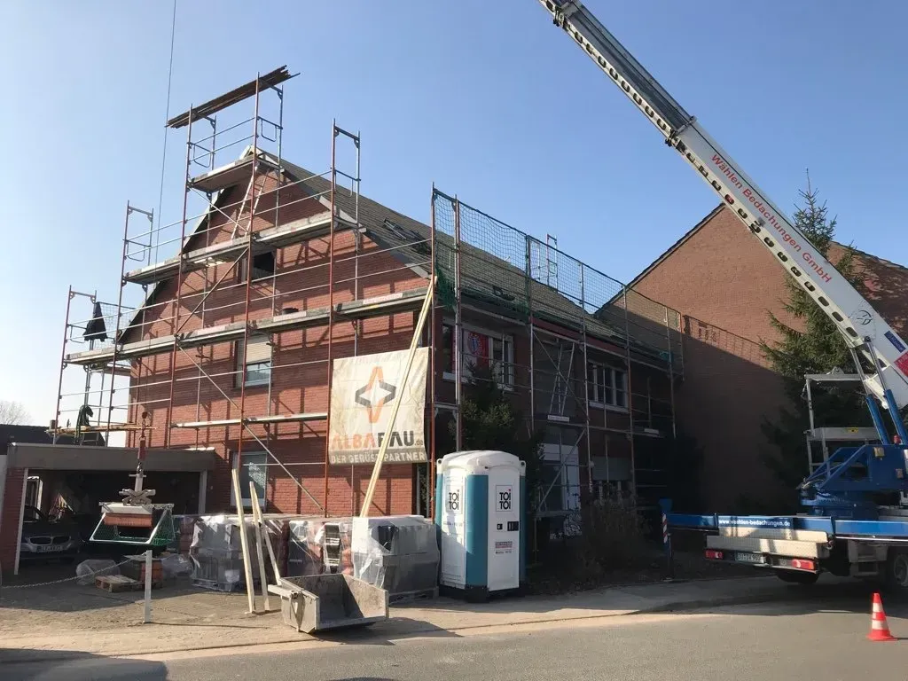 Ein Baugerüst umgibt ein rotes Gebäude; in der Nähe hebt ein Kran Baumaterialien an.