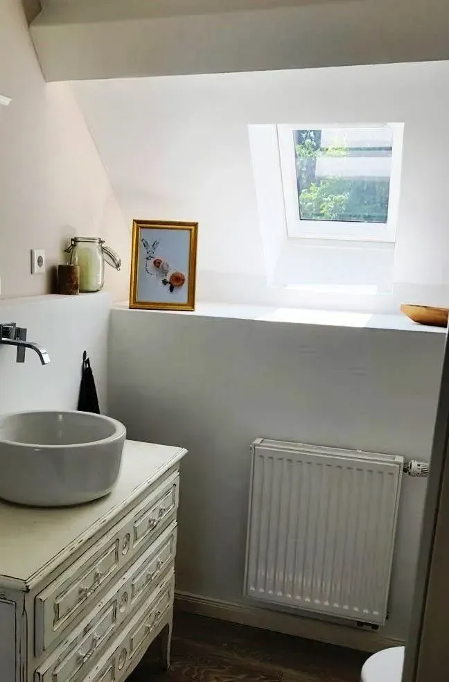 Badezimmer mit weißem Waschbecken, verziertem Waschtisch, Heizkörper und kleinem Fenster über einem Sims.
